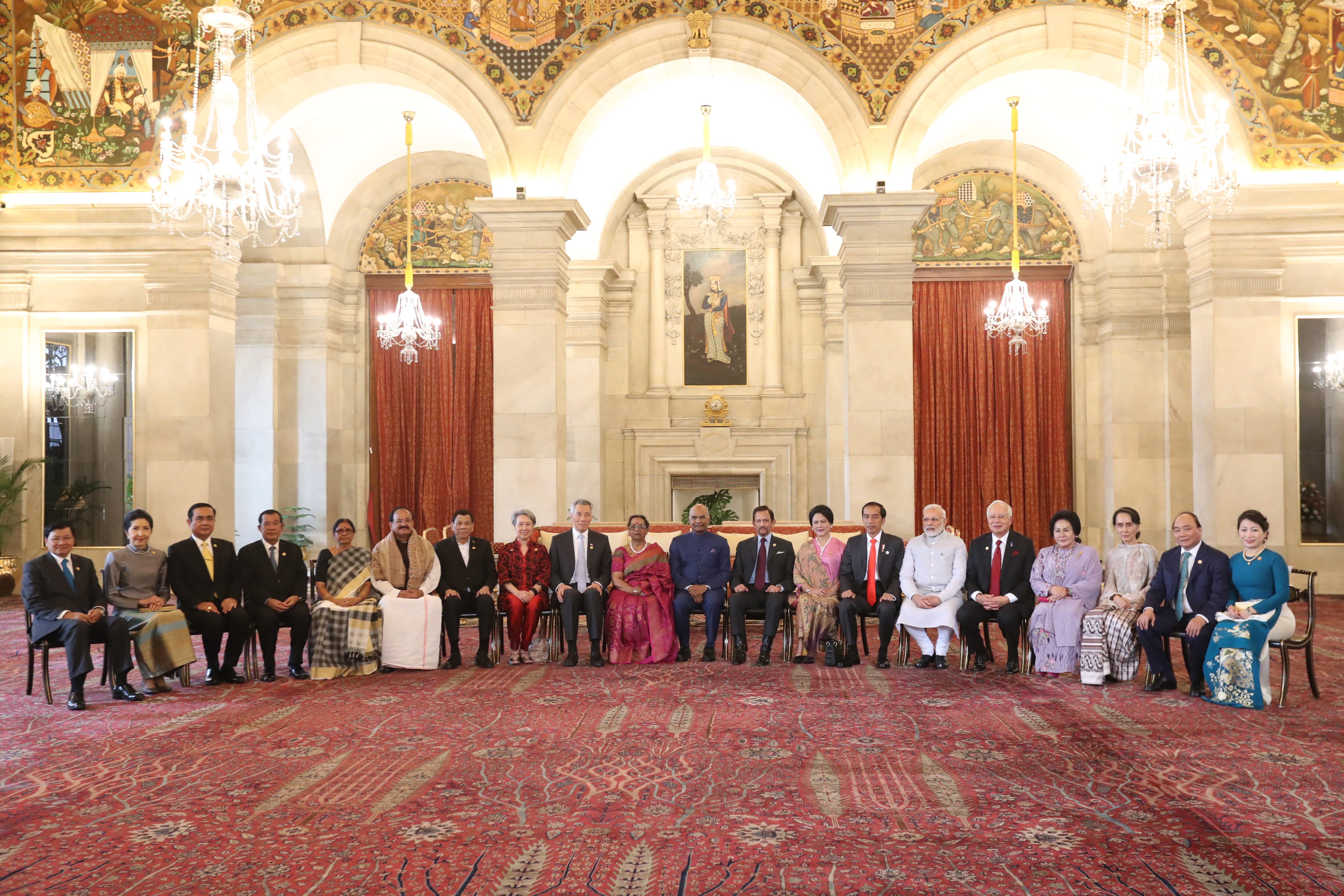 Group of people sitting in ornate hall with red patterned carpet and detailed wall decor.