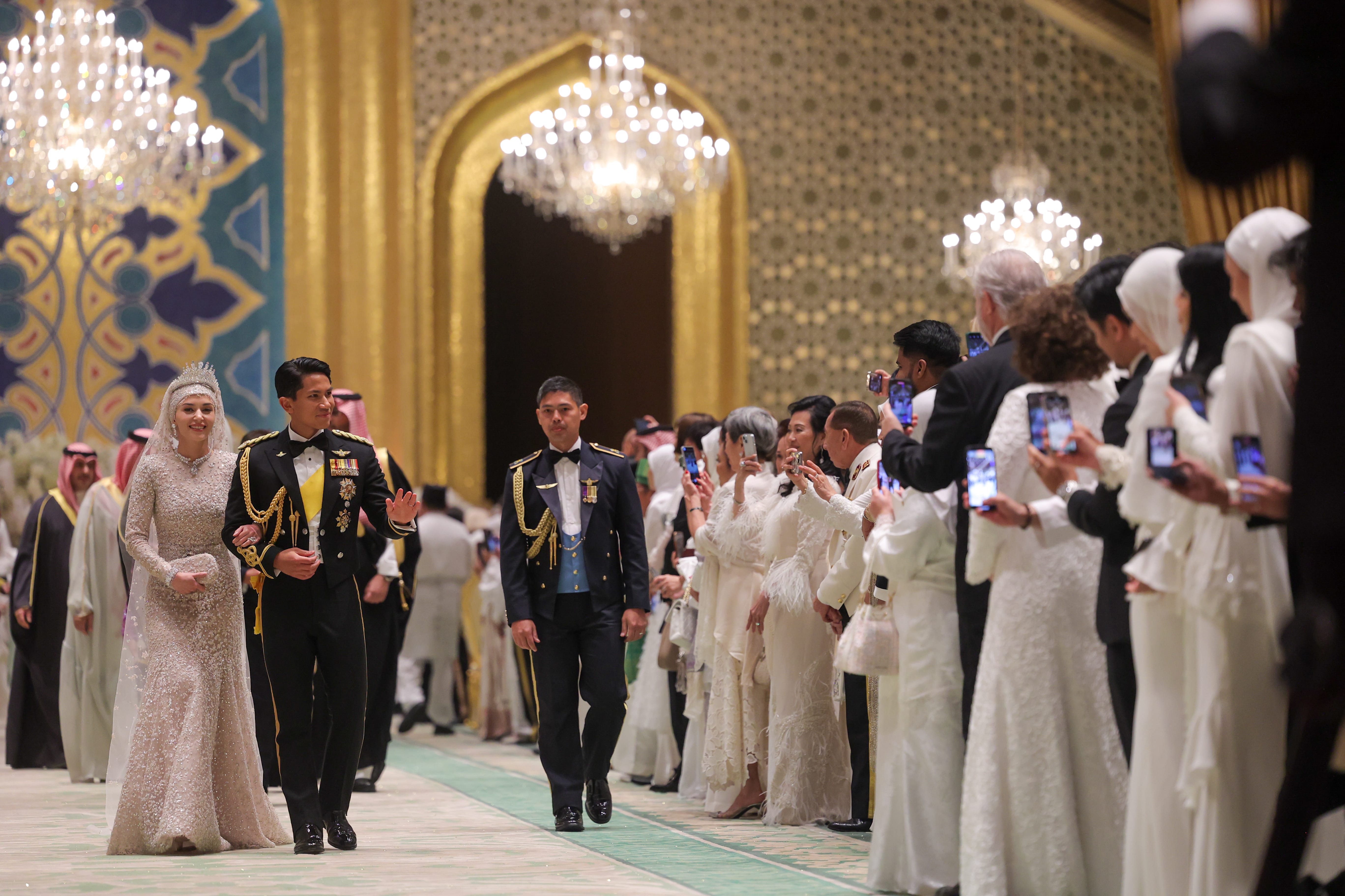 Bride in tiara, groom in military dress, walking by guests taking photos in decorated hall.