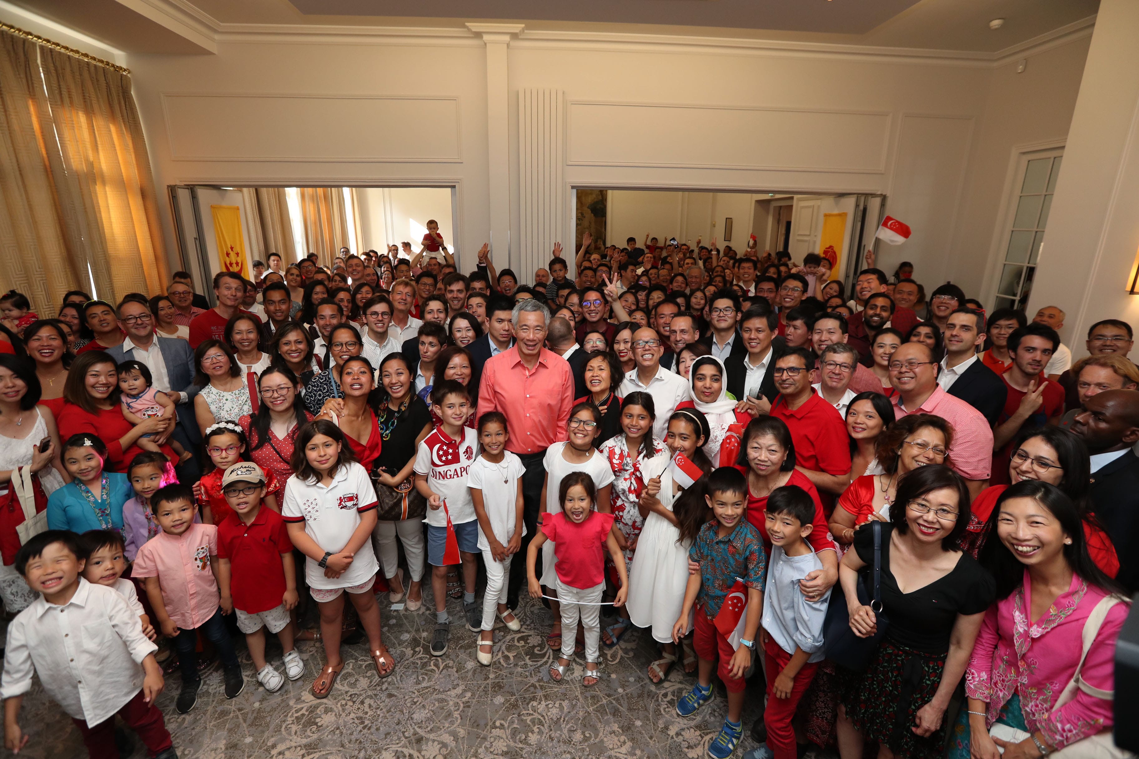 Large group of diverse people, including Lee Hsien Loong, pose indoors with some holding Singapore flags.