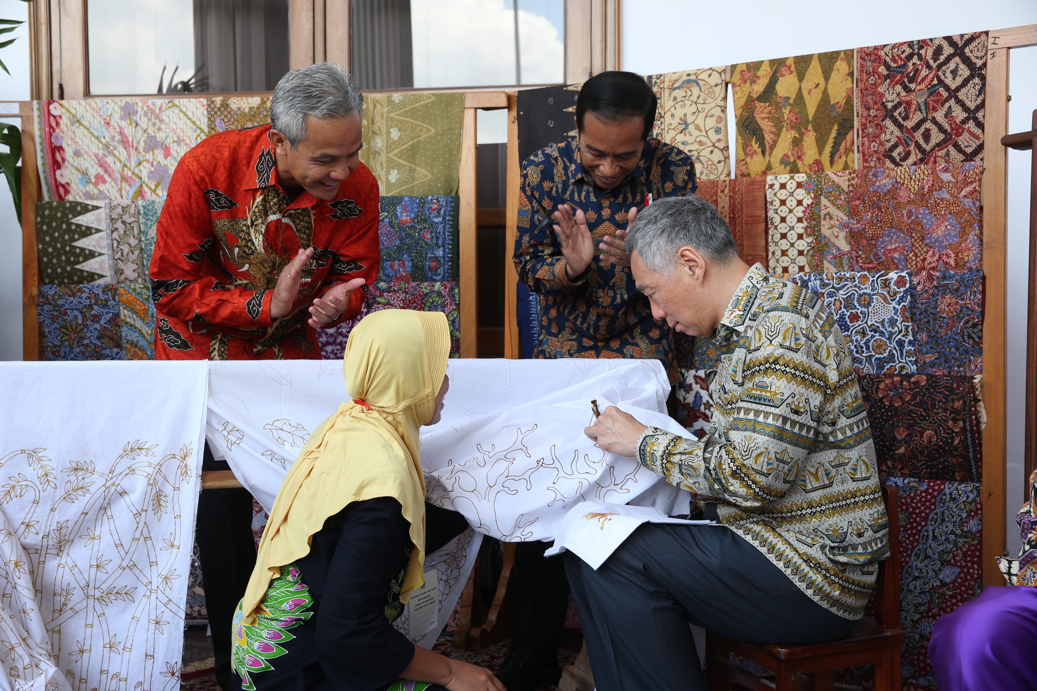 Three men and a woman working on batik cloths in front of batik display cloths.