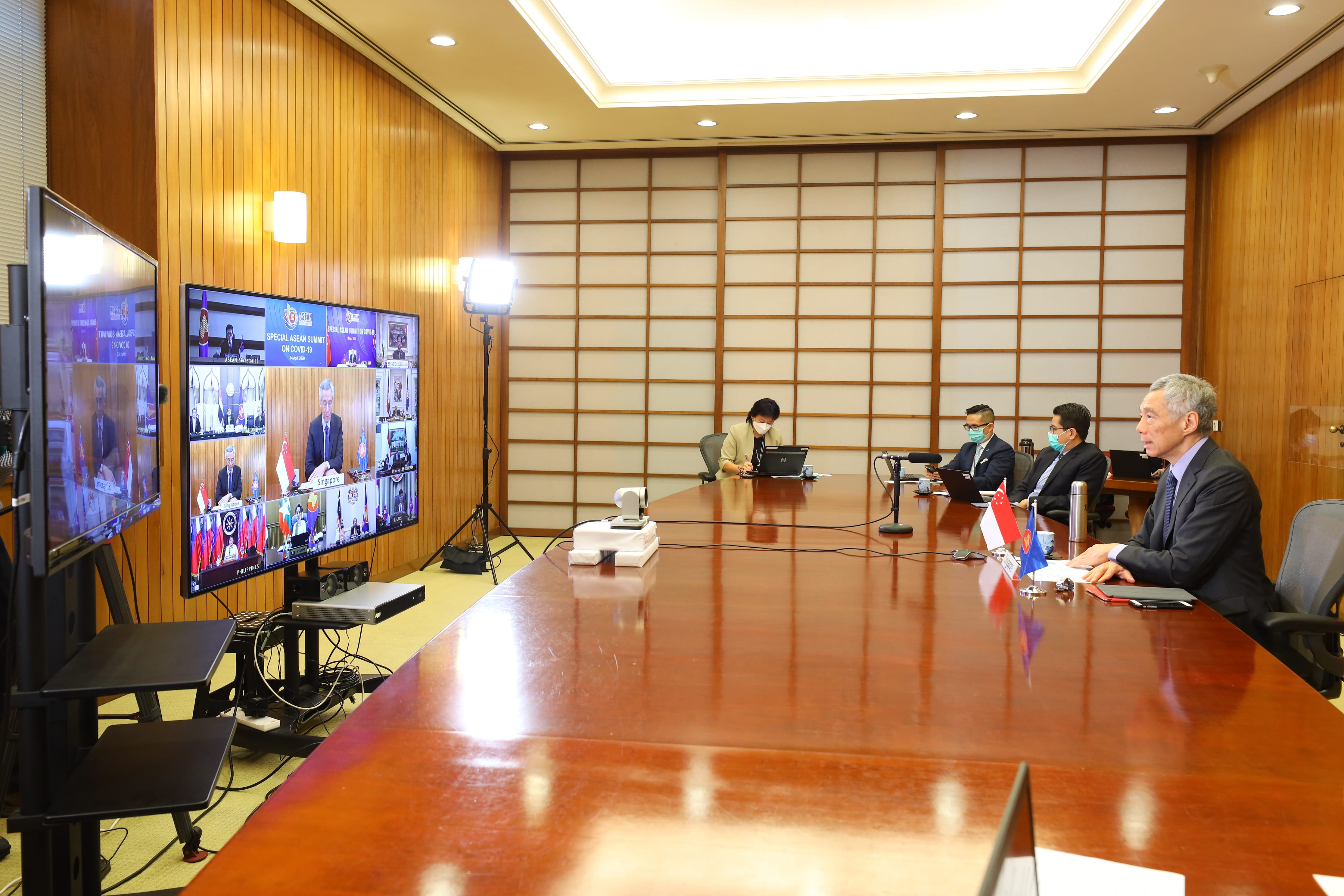Conference room with people at a long table; video screen displays "ASEAN Summit on COVID-19".