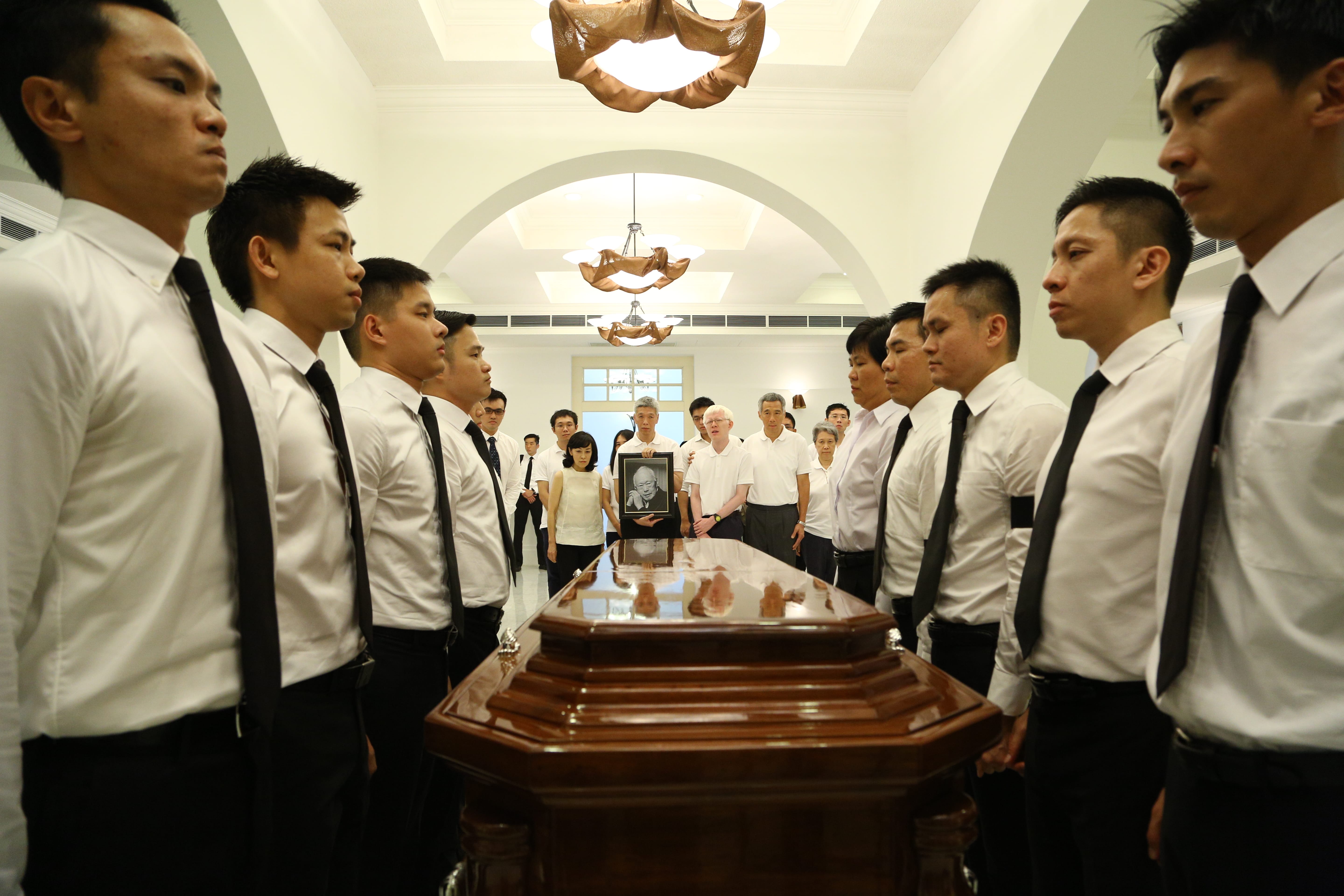 Guards in white shirts flank a wooden casket; a framed photo of Lee Kuan Yew visible.