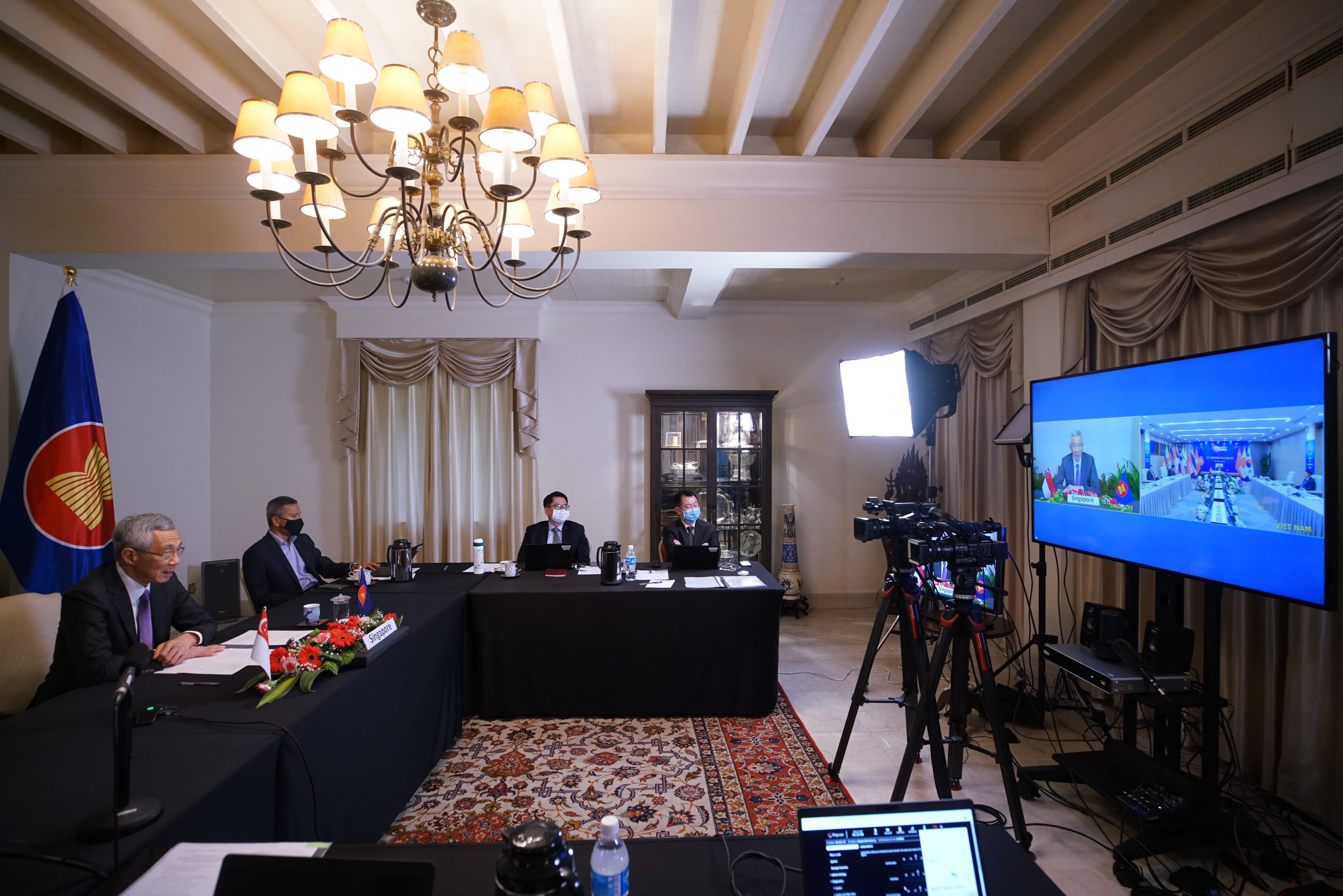People in suits at a long conference table are filmed for a video conference displayed on a large screen.