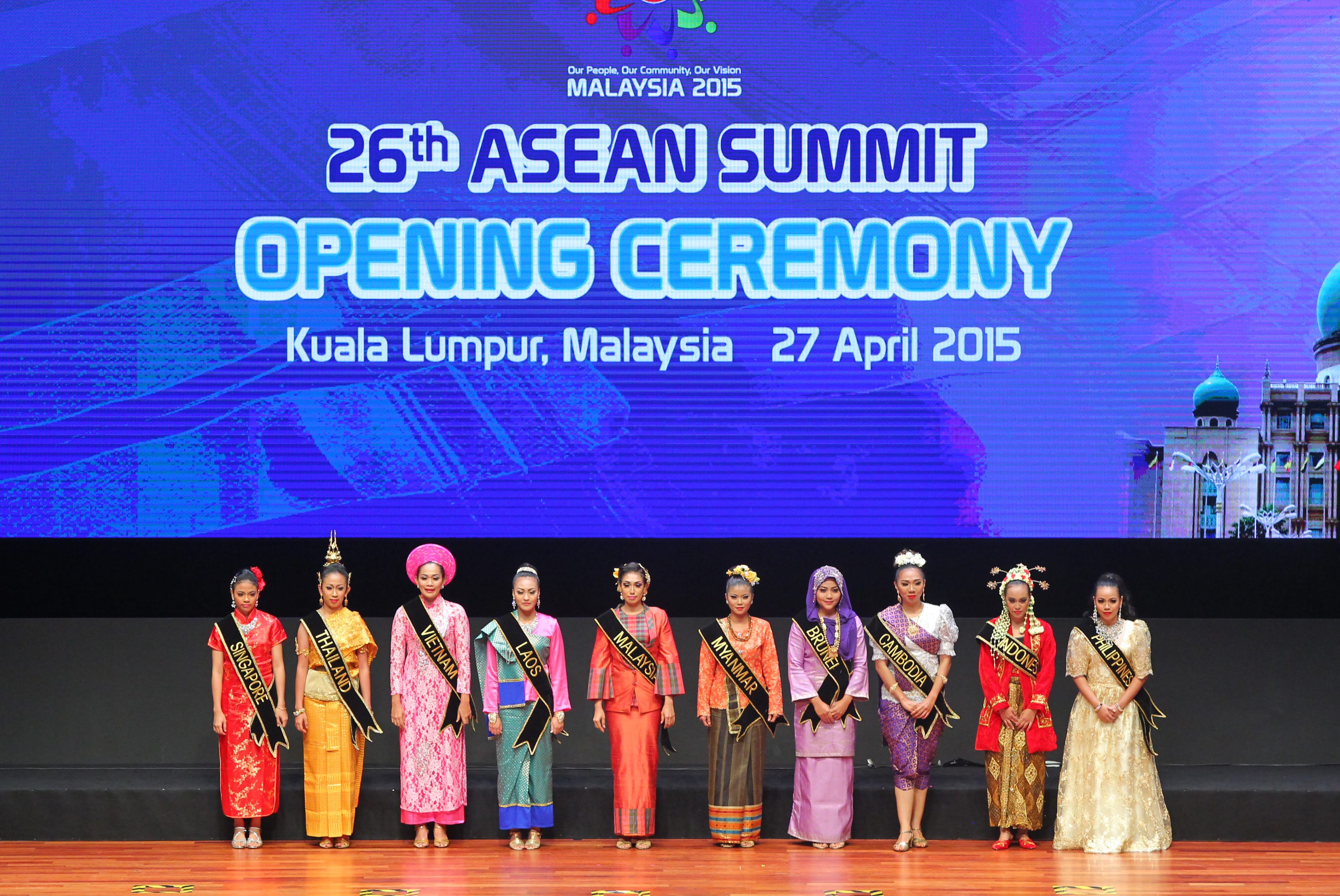 Eleven women on stage in formalwear with country sashes, facing front, "26th ASEAN Summit" backdrop.