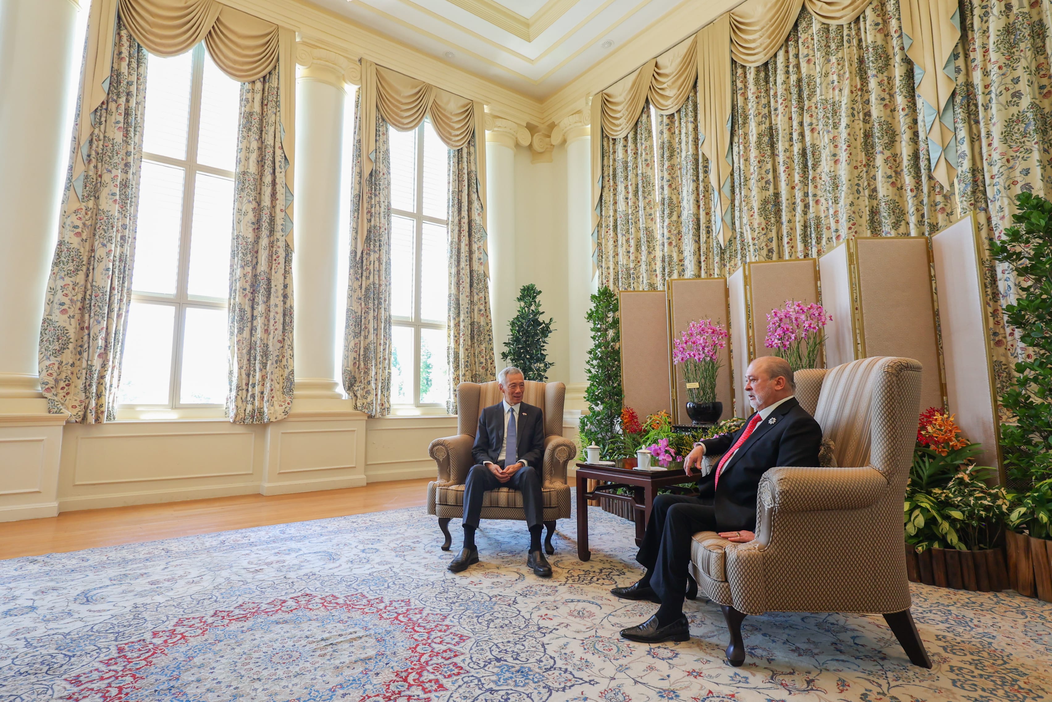 Lee Hsien Loong and another man sit in chairs in a room with large windows and floral curtains.