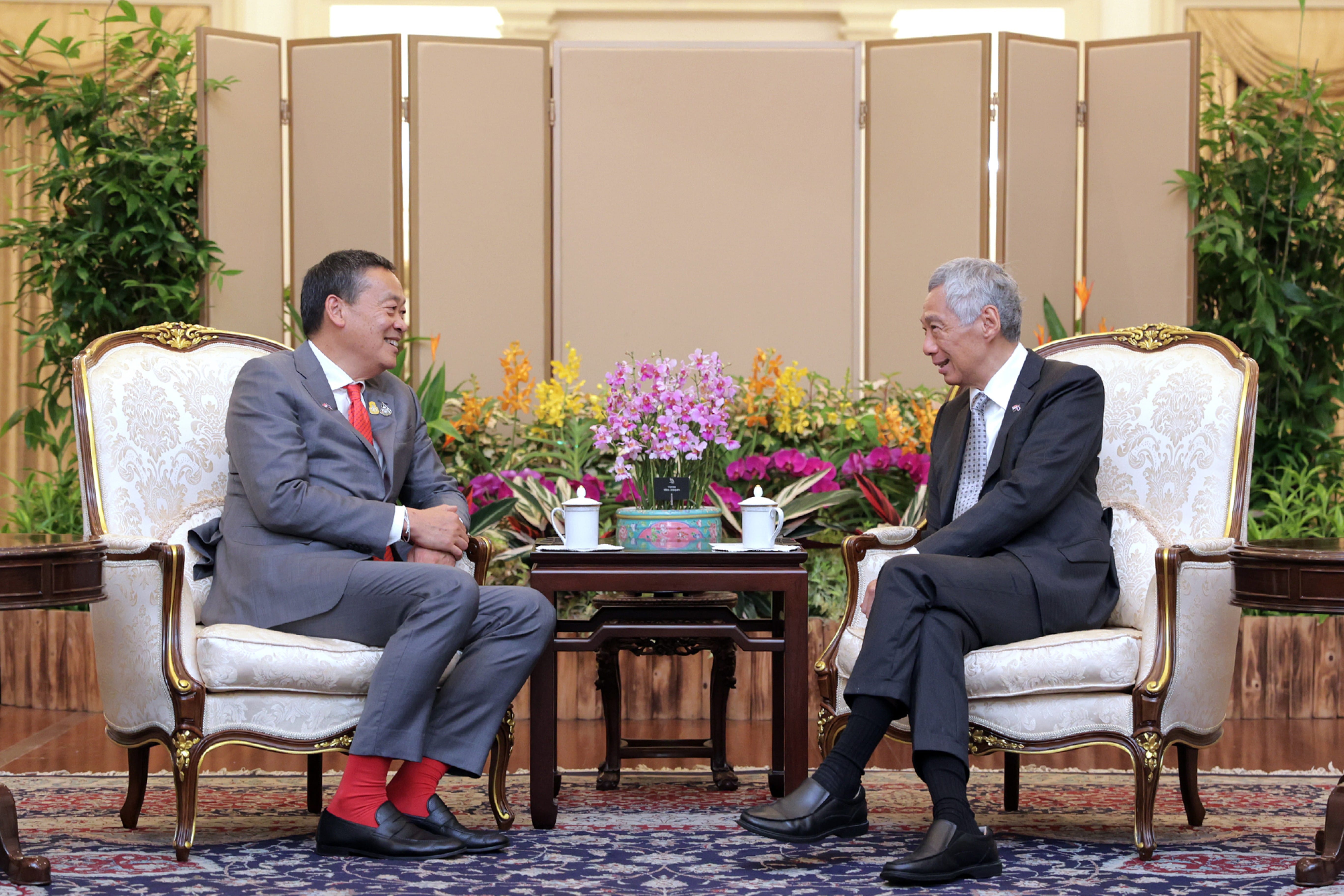 Two men in suits sit in ornate chairs facing each other across a small table with flowers and tea cups.