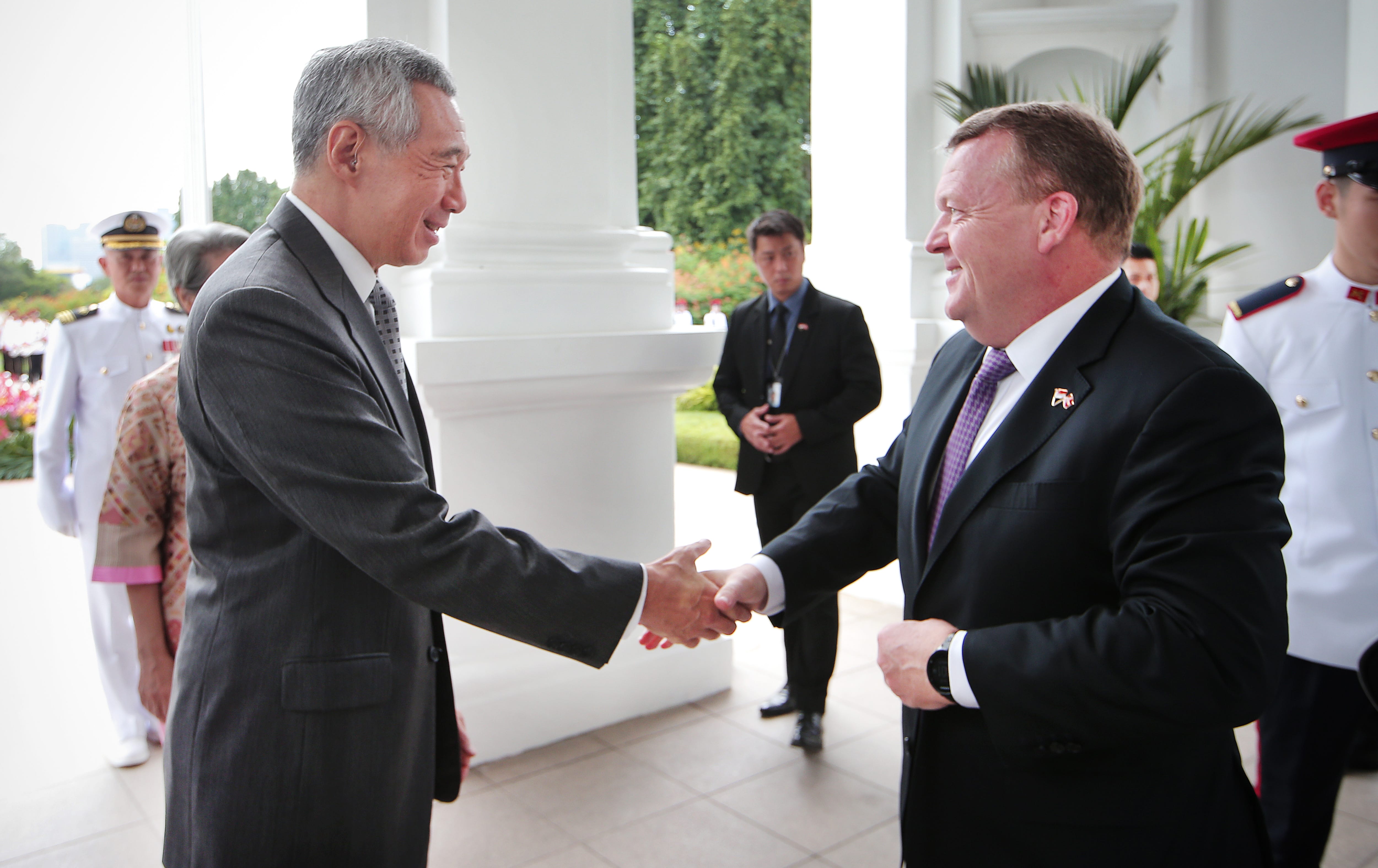 Two men in suits shake hands outdoors. Staff members in uniform stand in the background.
