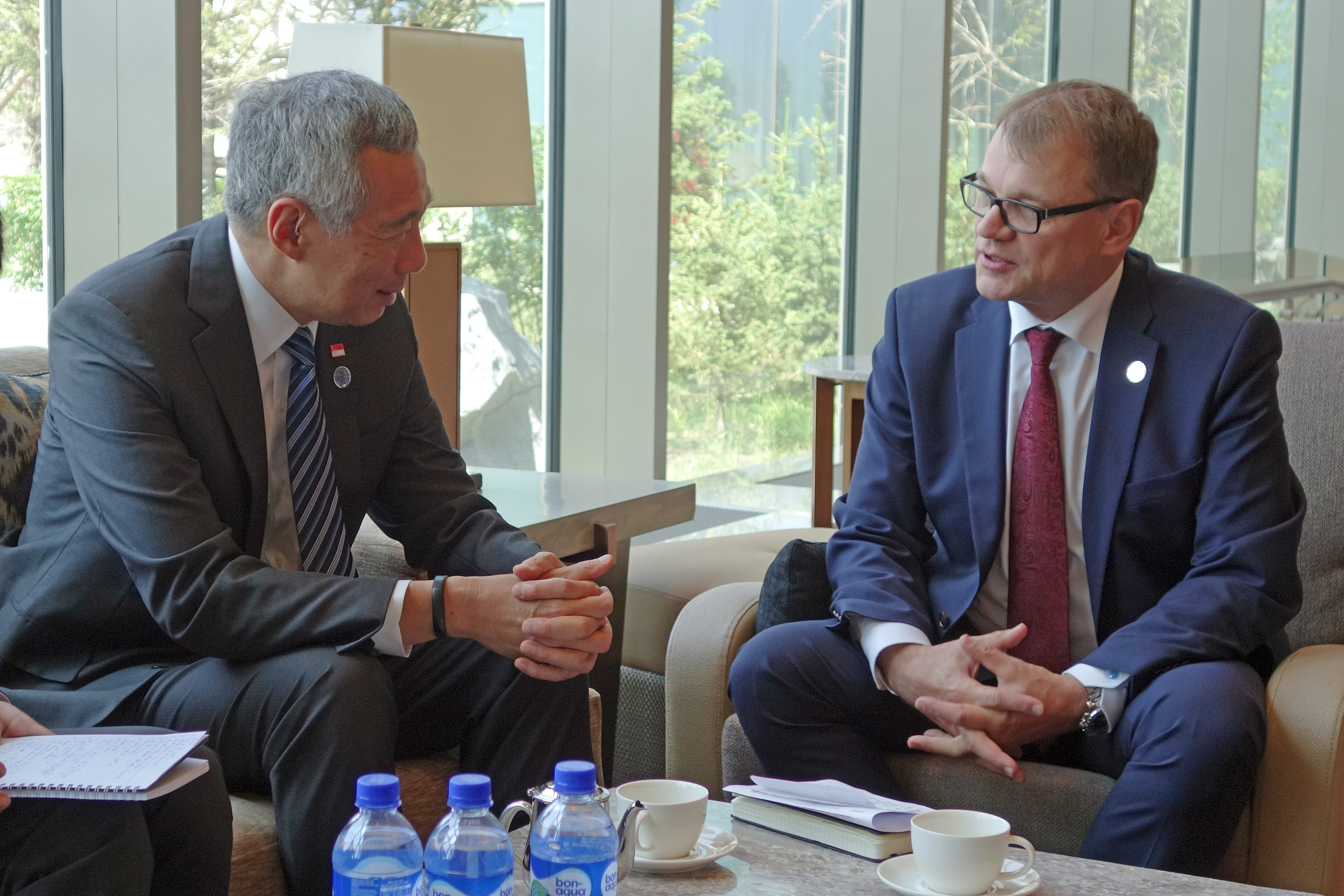 Two men in suits seated, face-to-face, near Bon Aqua bottles and teacups by a window.