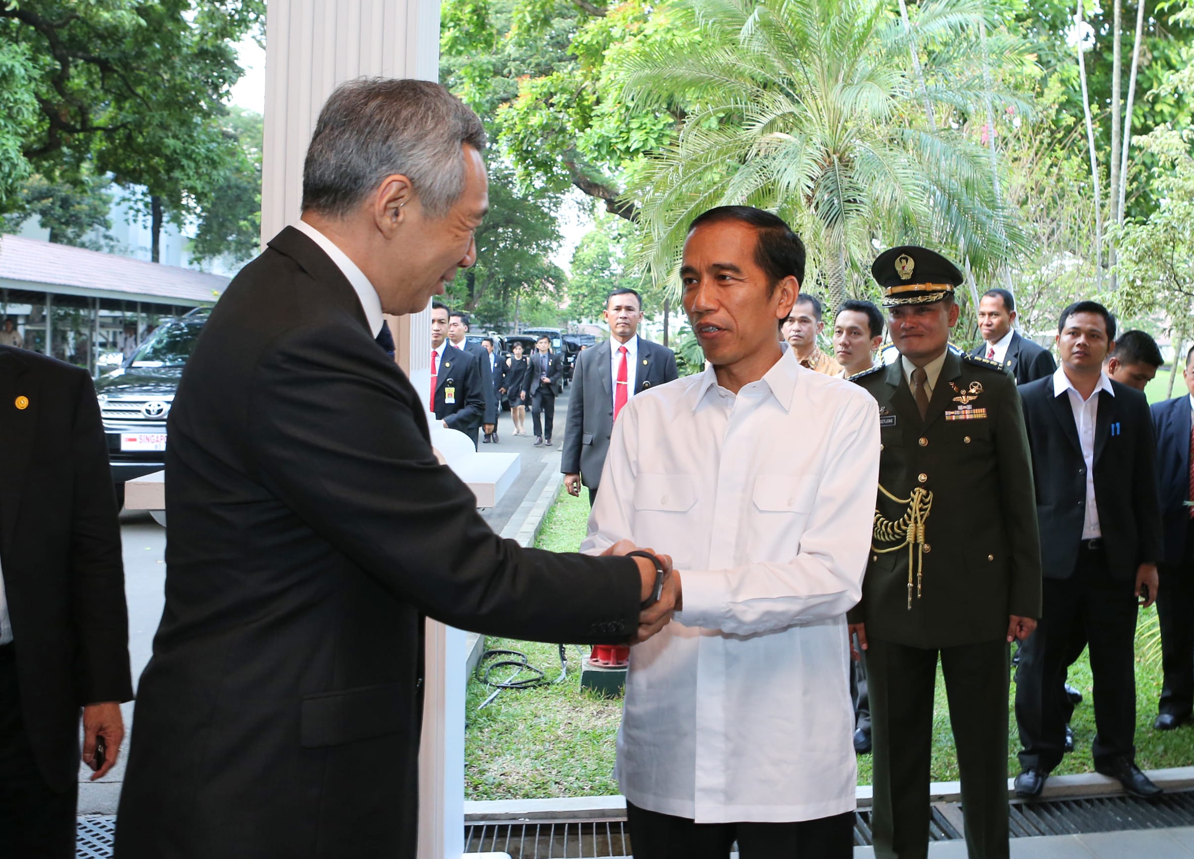 Lee Hsien Loong shakes hands with Jokowi amid security personnel and outdoor greenery.