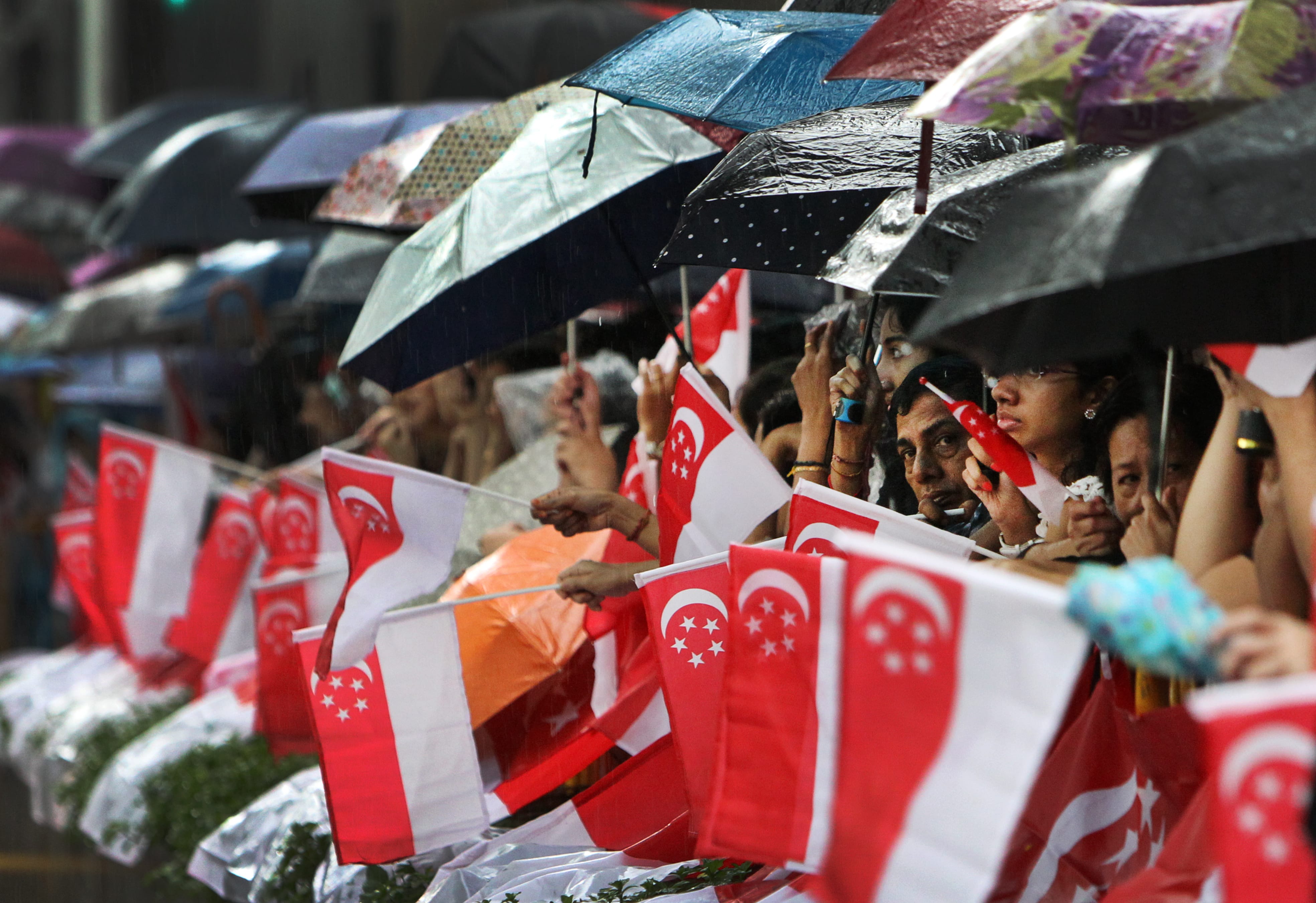 People under umbrellas wave Singapore flags in the rain.