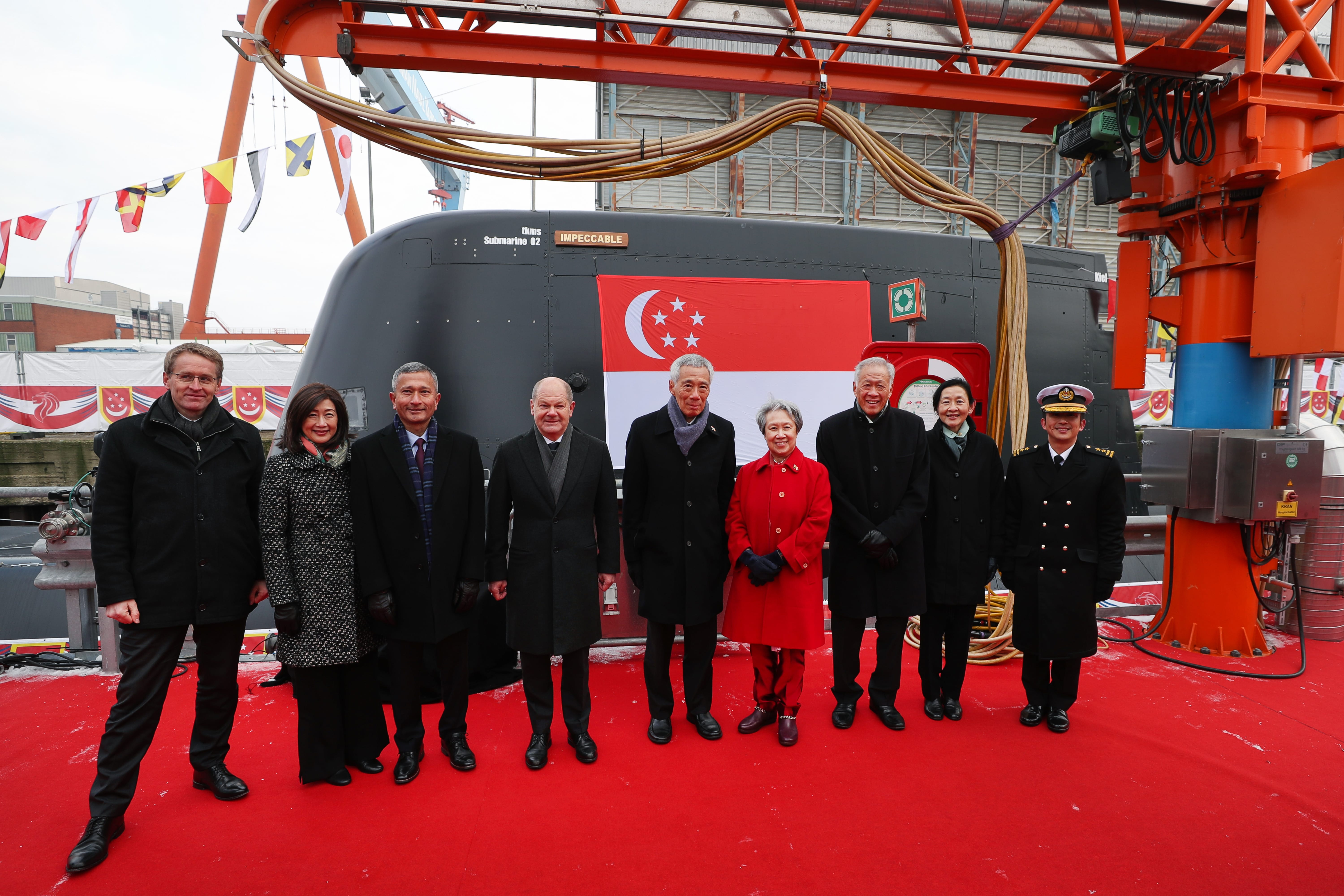 Group of nine people on red carpet in front of dark submarine with Singapore flag.