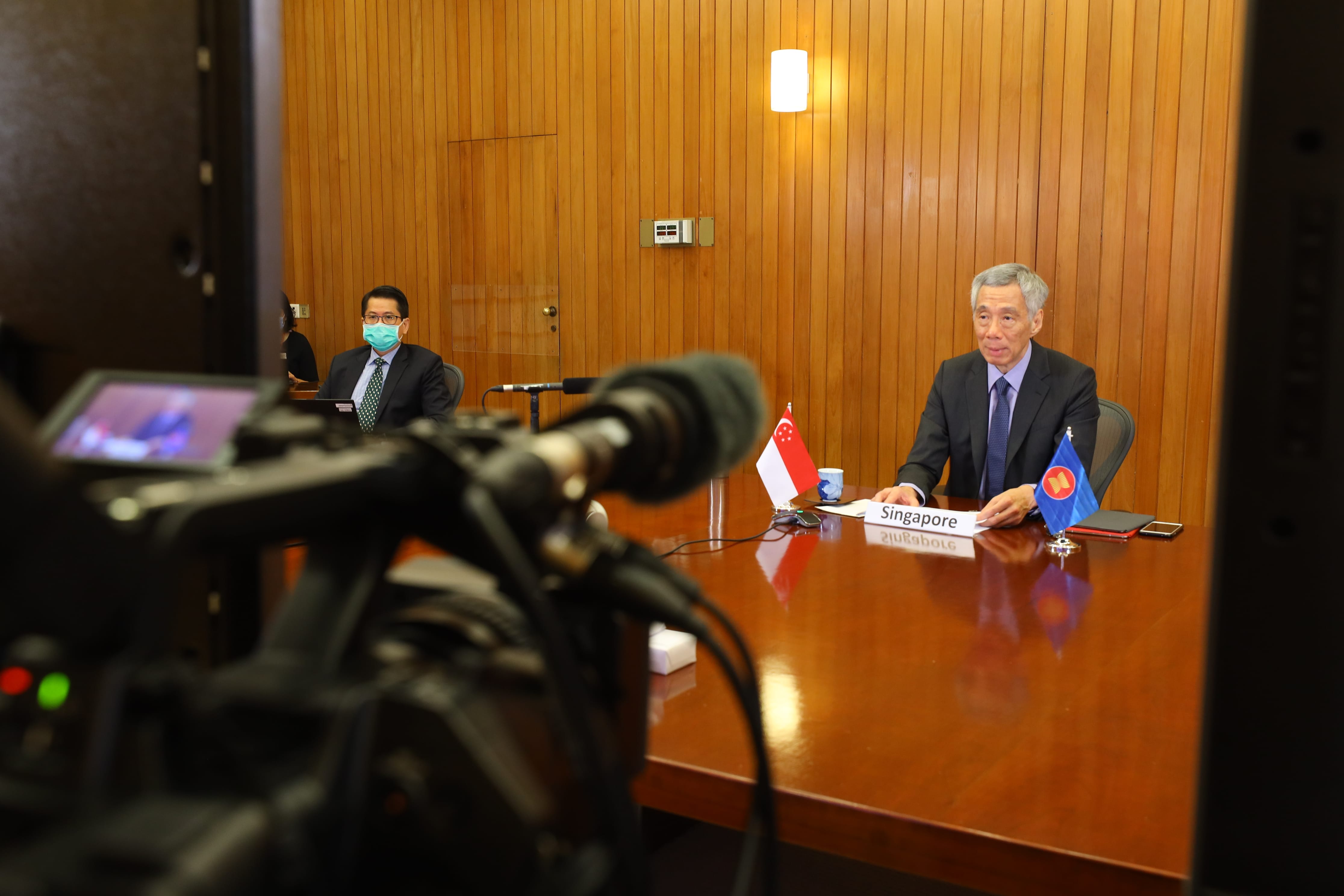 Two men at a wooden table with country flags, being filmed. Lee Hsien Loong is visible.