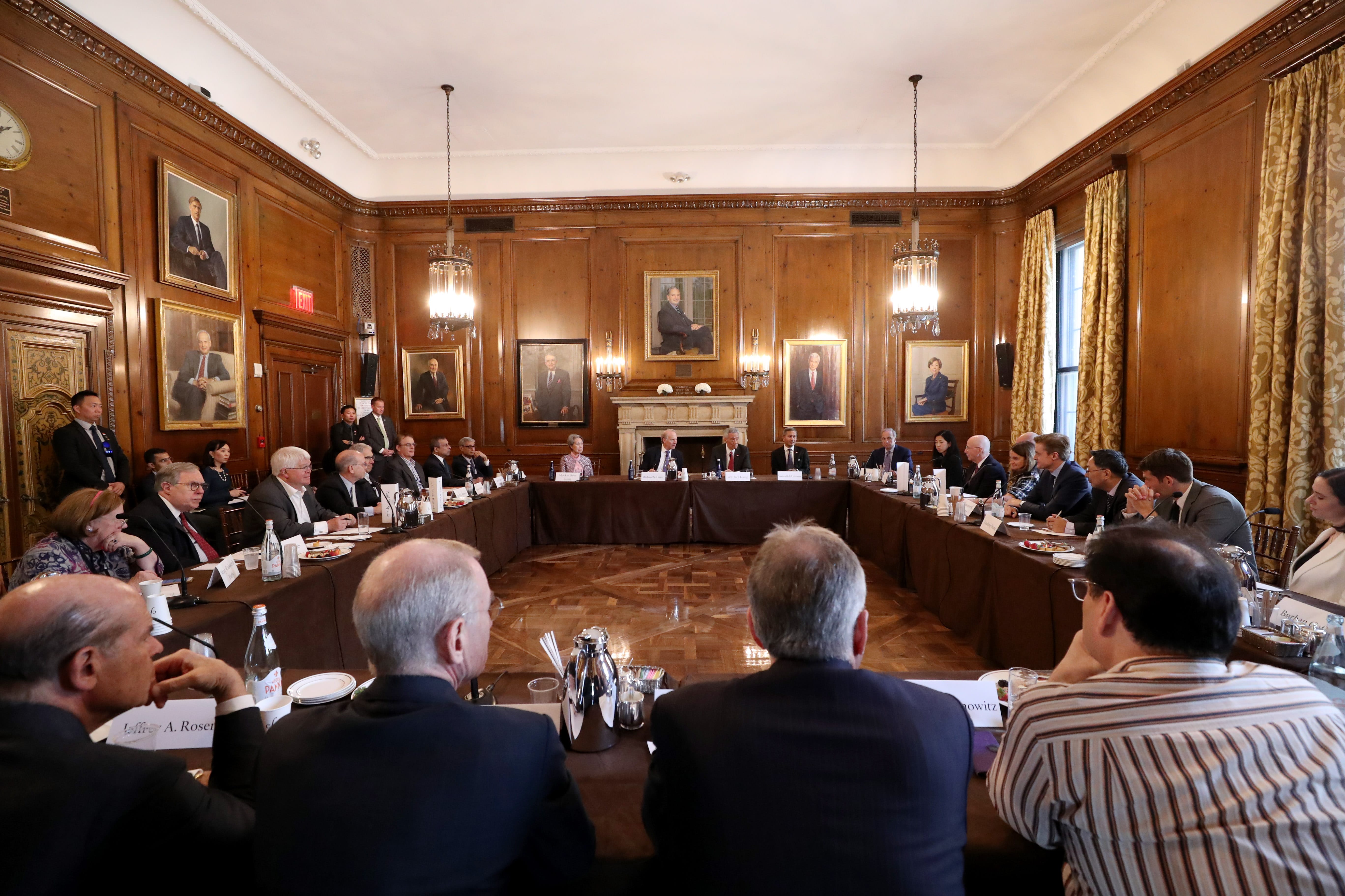 People at long U-shaped table in wood-paneled room, portraits on wall.