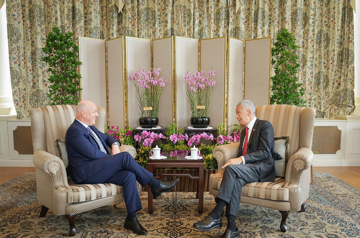 Two men in suits converse while seated in armchairs, with a tea table and floral backdrop.