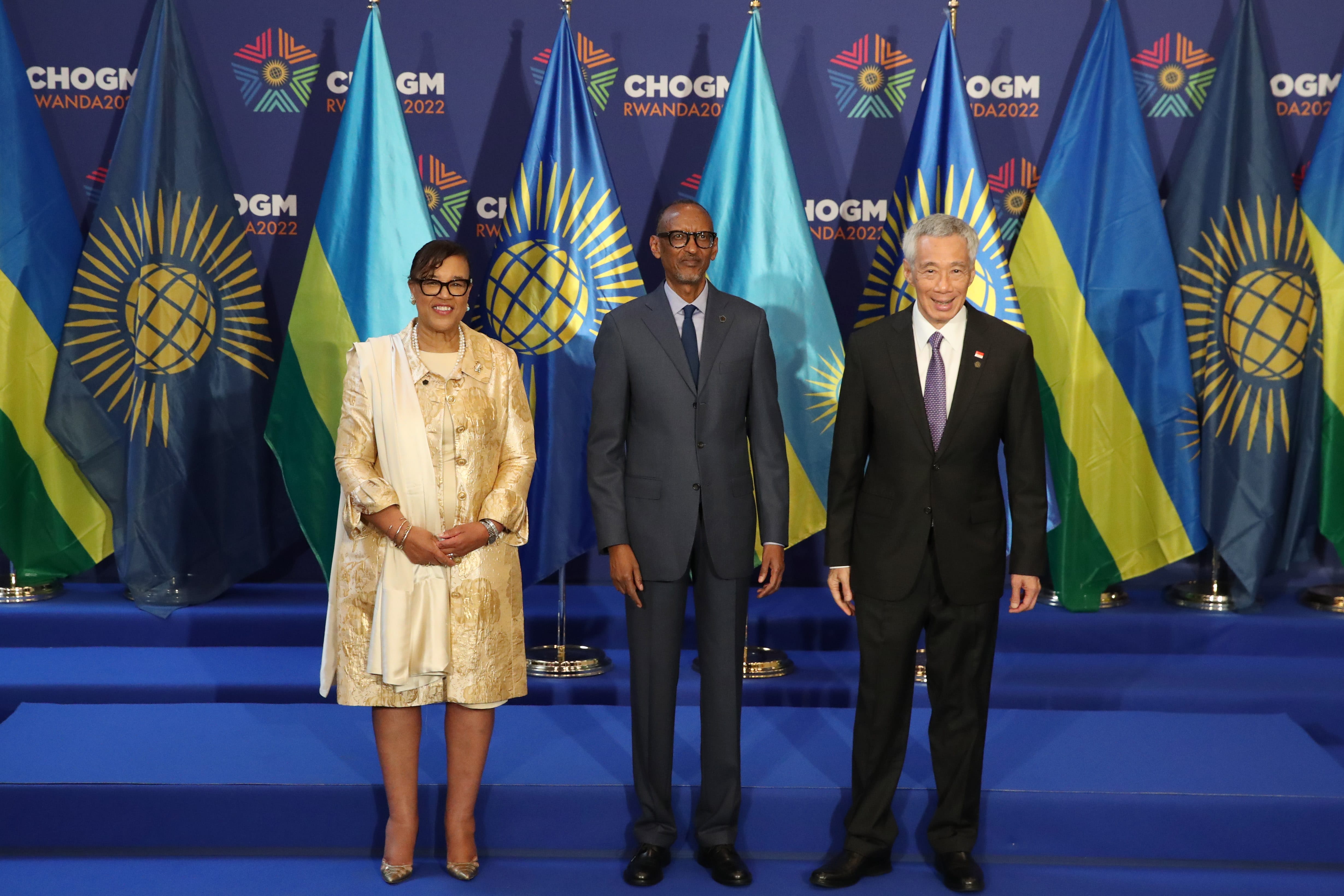 Three people stand before flags with "CHOGM" and globe graphics. Each person wears business attire.