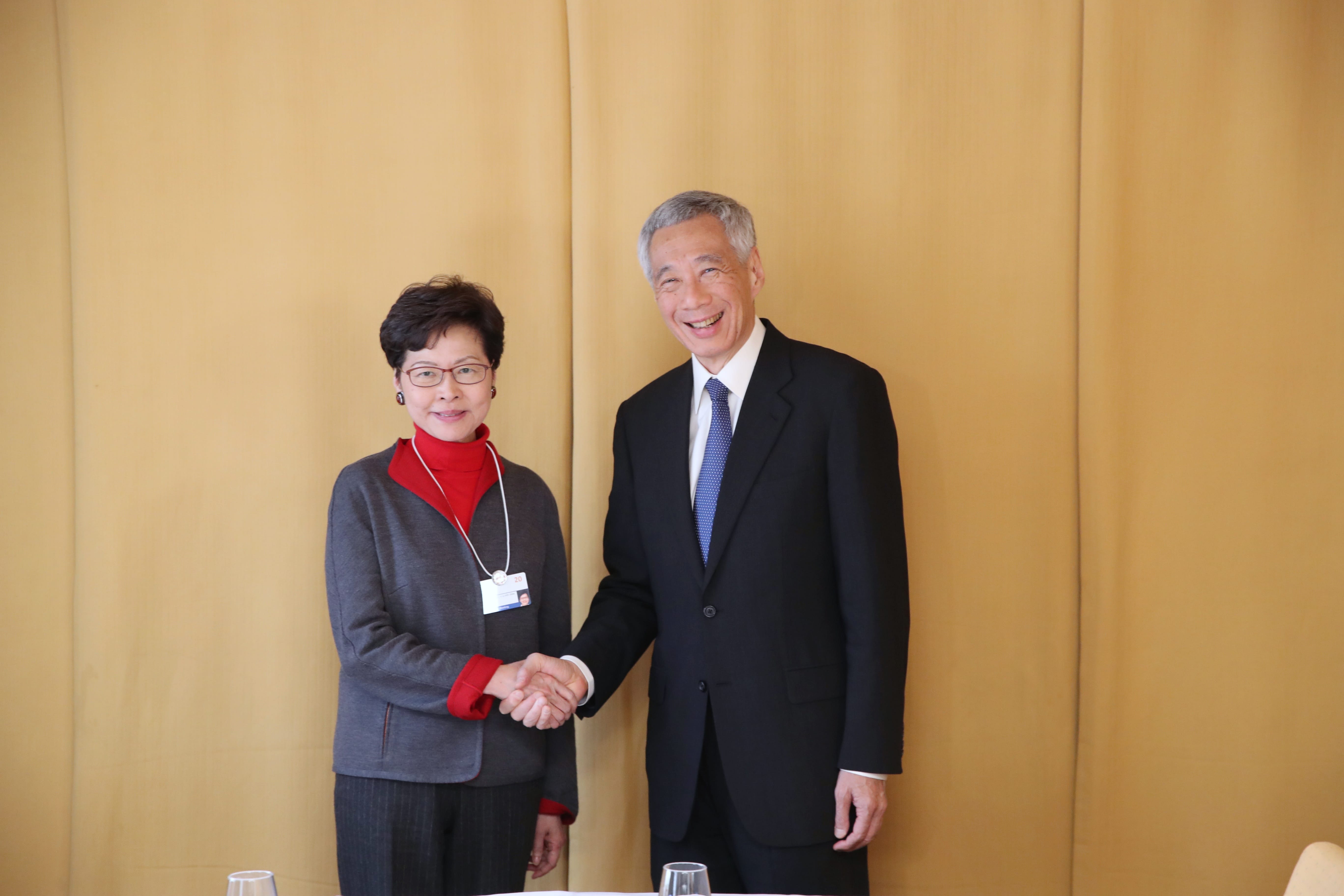Lee Hsien Loong shaking hands with a woman in glasses and red sweater against a gold background.