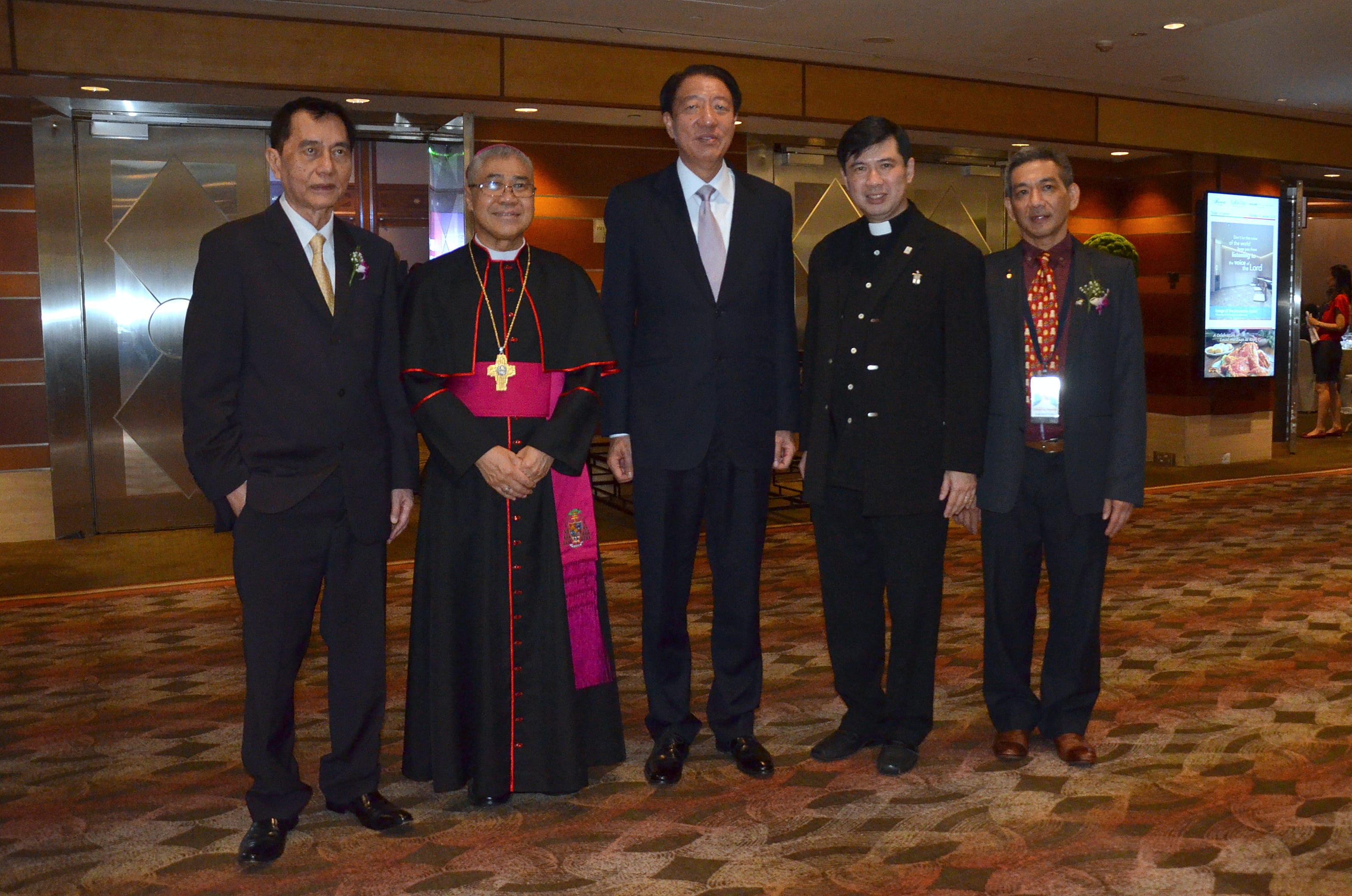 Five men in formal wear, including a bishop and a priest, stand in a hotel-like room.