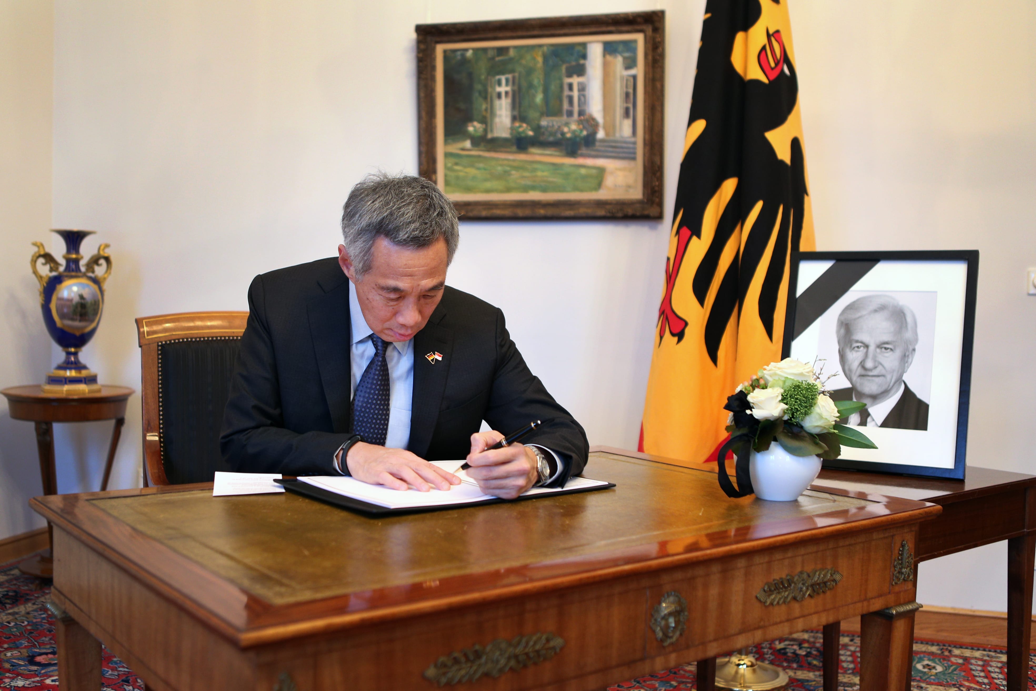 Lee Hsien Loong writing at a desk with a German flag, picture of Lee Kuan Yew, and flowers nearby.