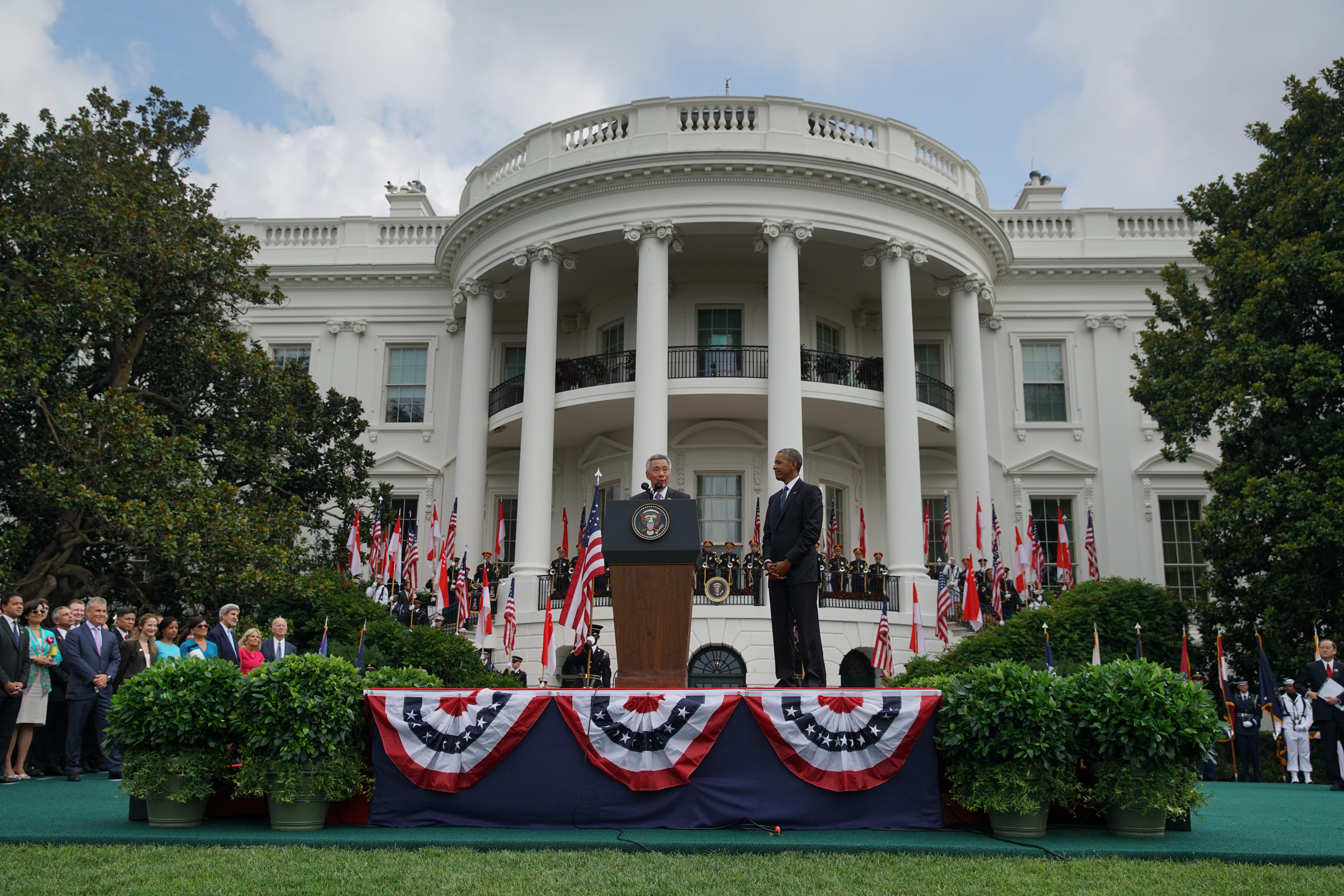 White House lawn event: speaker at podium, Obama stands nearby, flags, and uniformed honor guard.