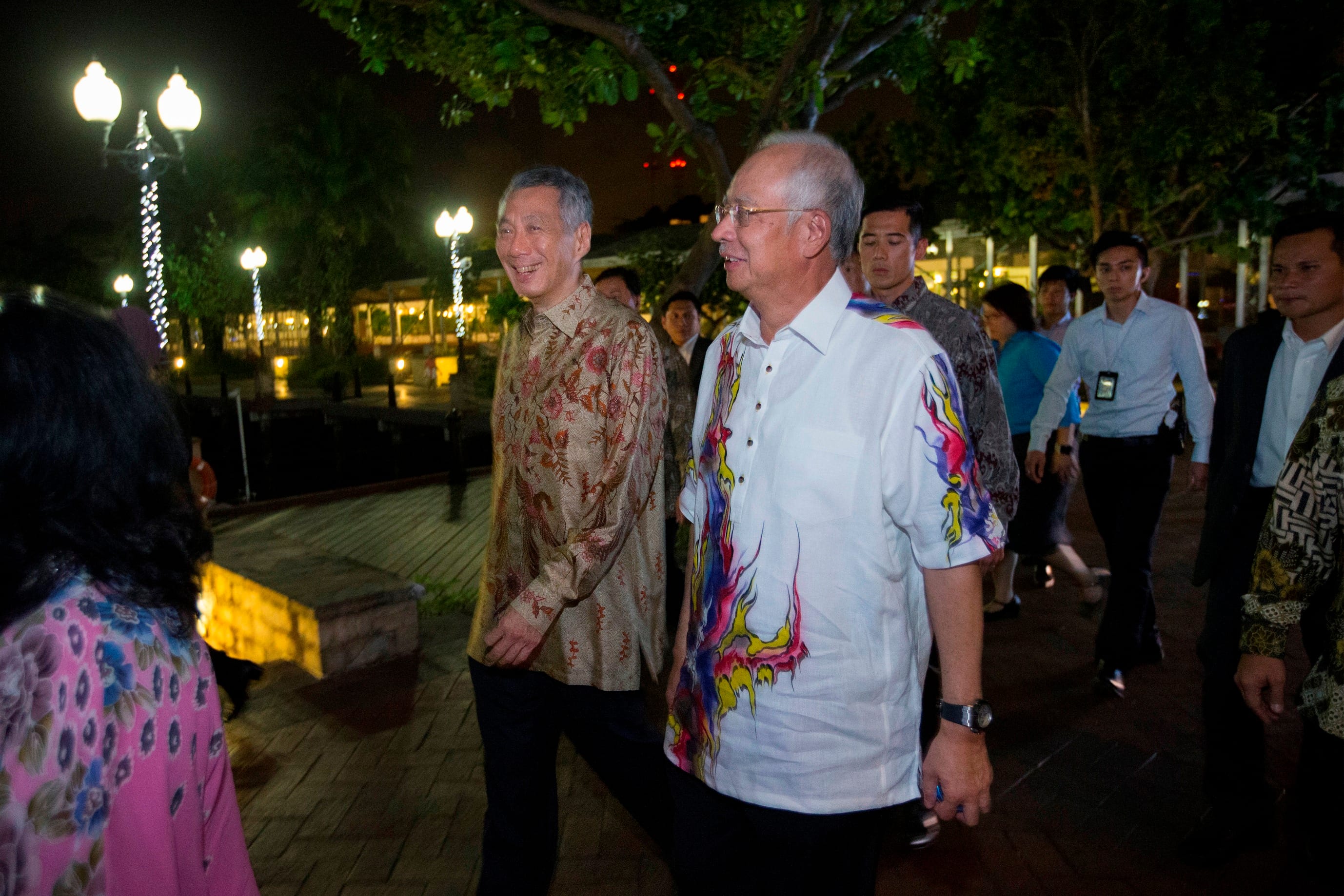 Lee Hsien Loong and another man walk on a path among others at night near trees and lights.