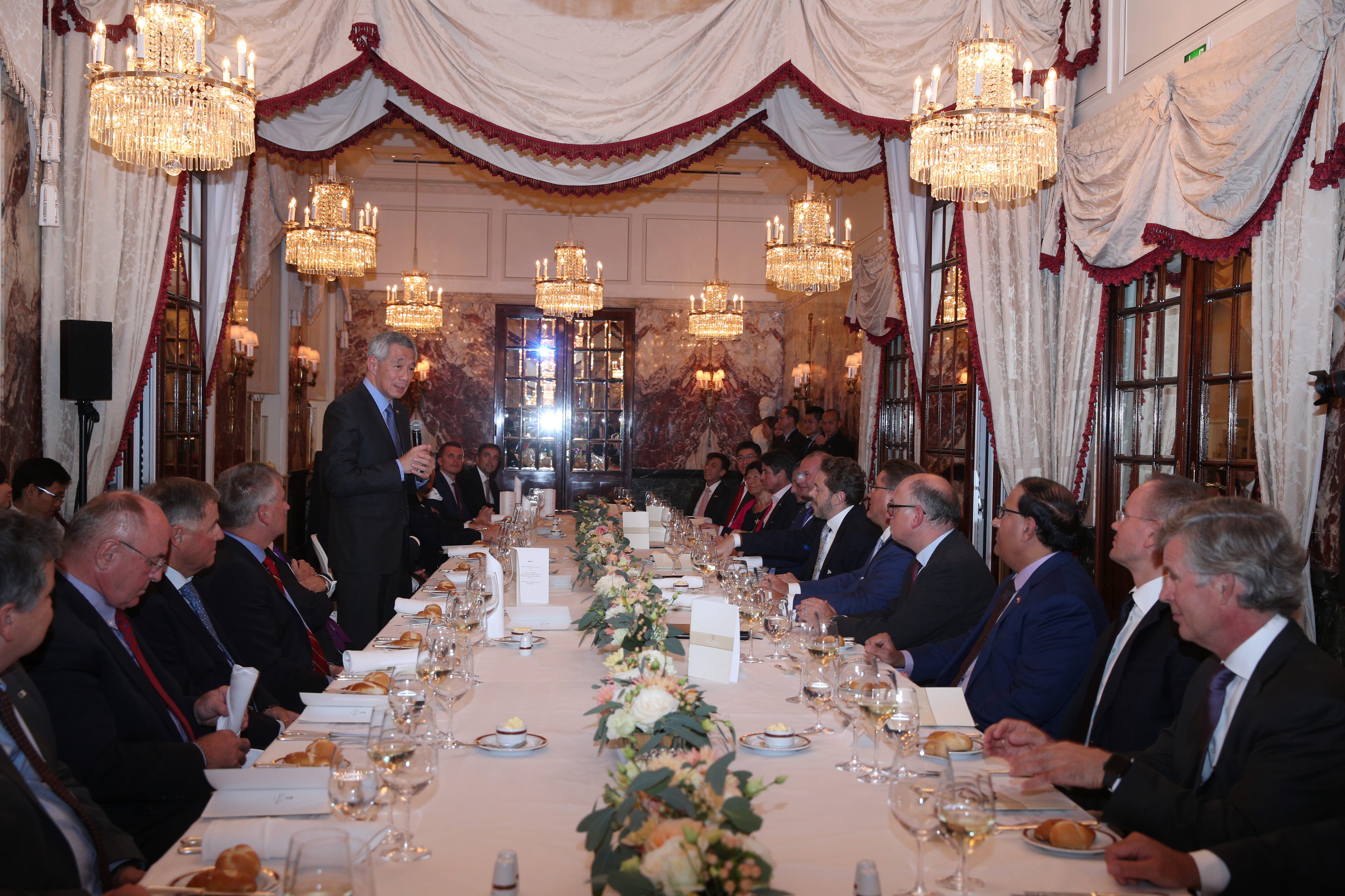 Lee Hsien Loong at a formal dinner, standing with a microphone, addressing people seated at long decorated table.