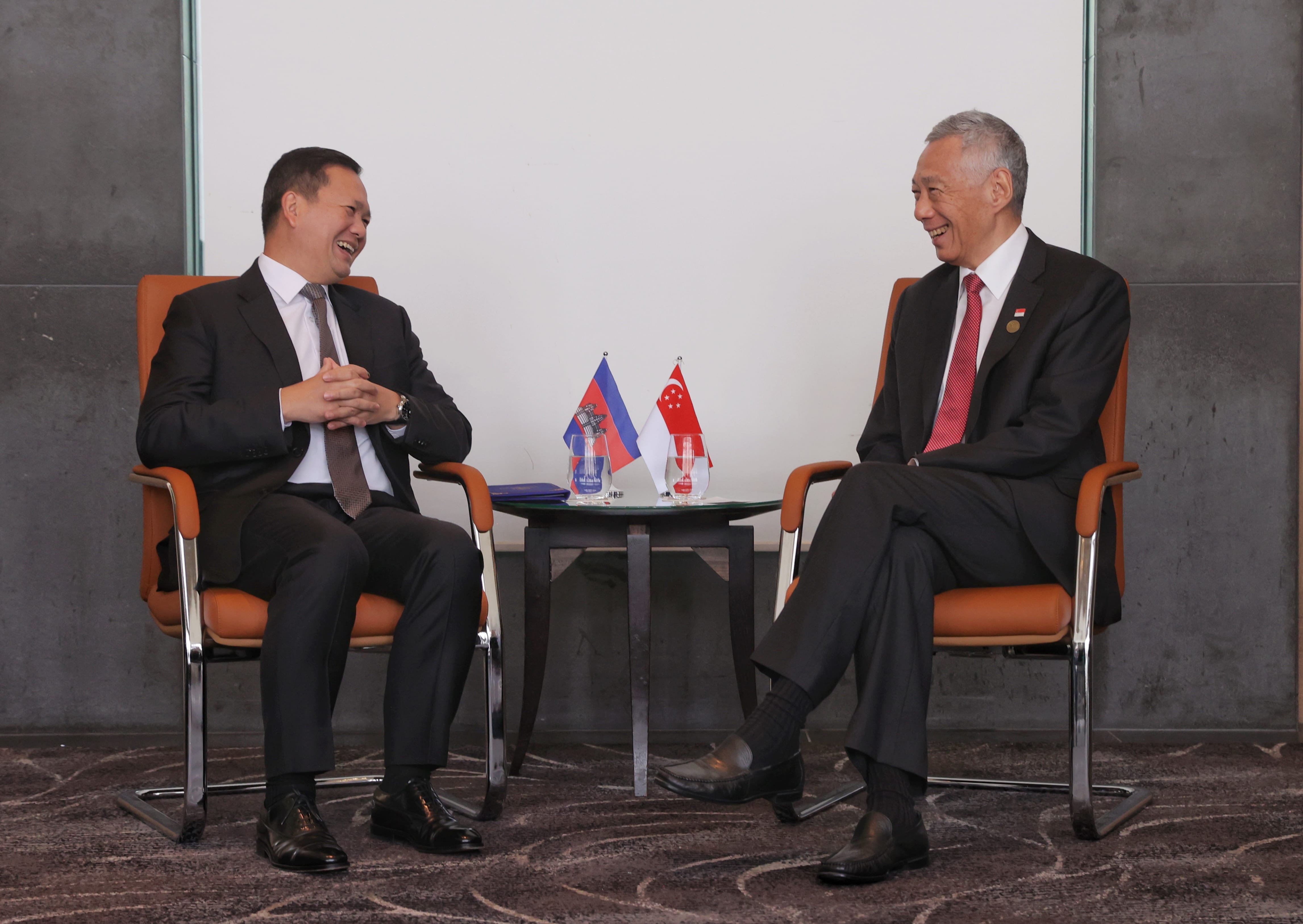 Two men in suits face each other, seated by Cambodia and Singapore flags.