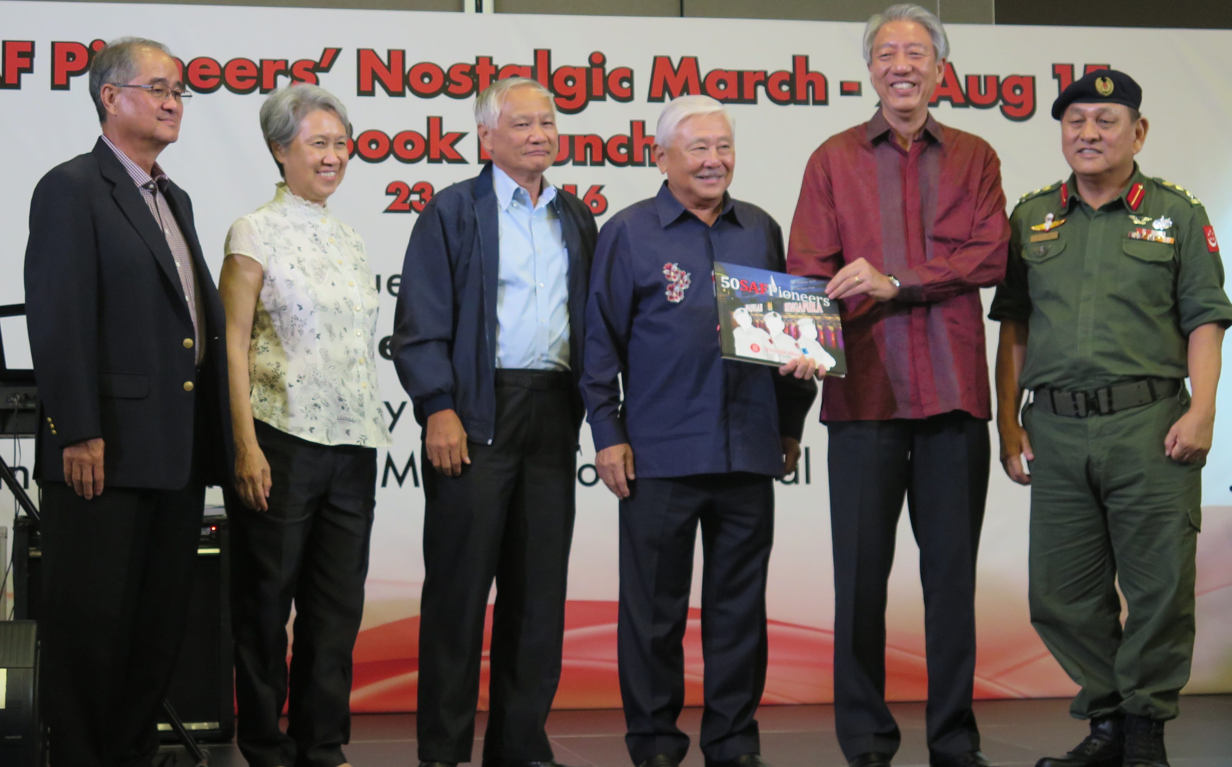 Six people stand together, one holding "50 SAF Pioneers" book; the last in military uniform.