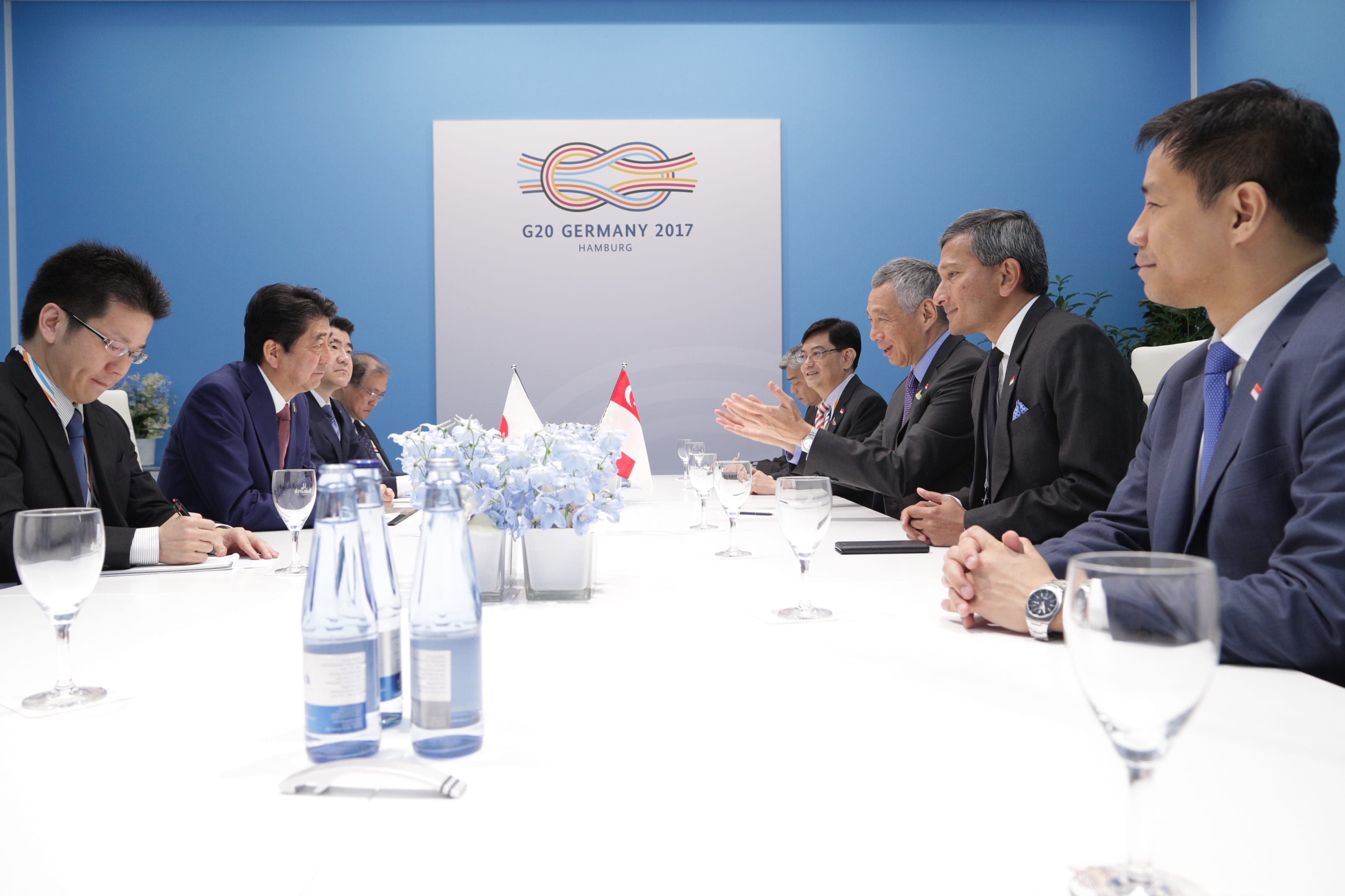 People in suits seated at a G20 Germany meeting with Japanese and Singaporean flags.