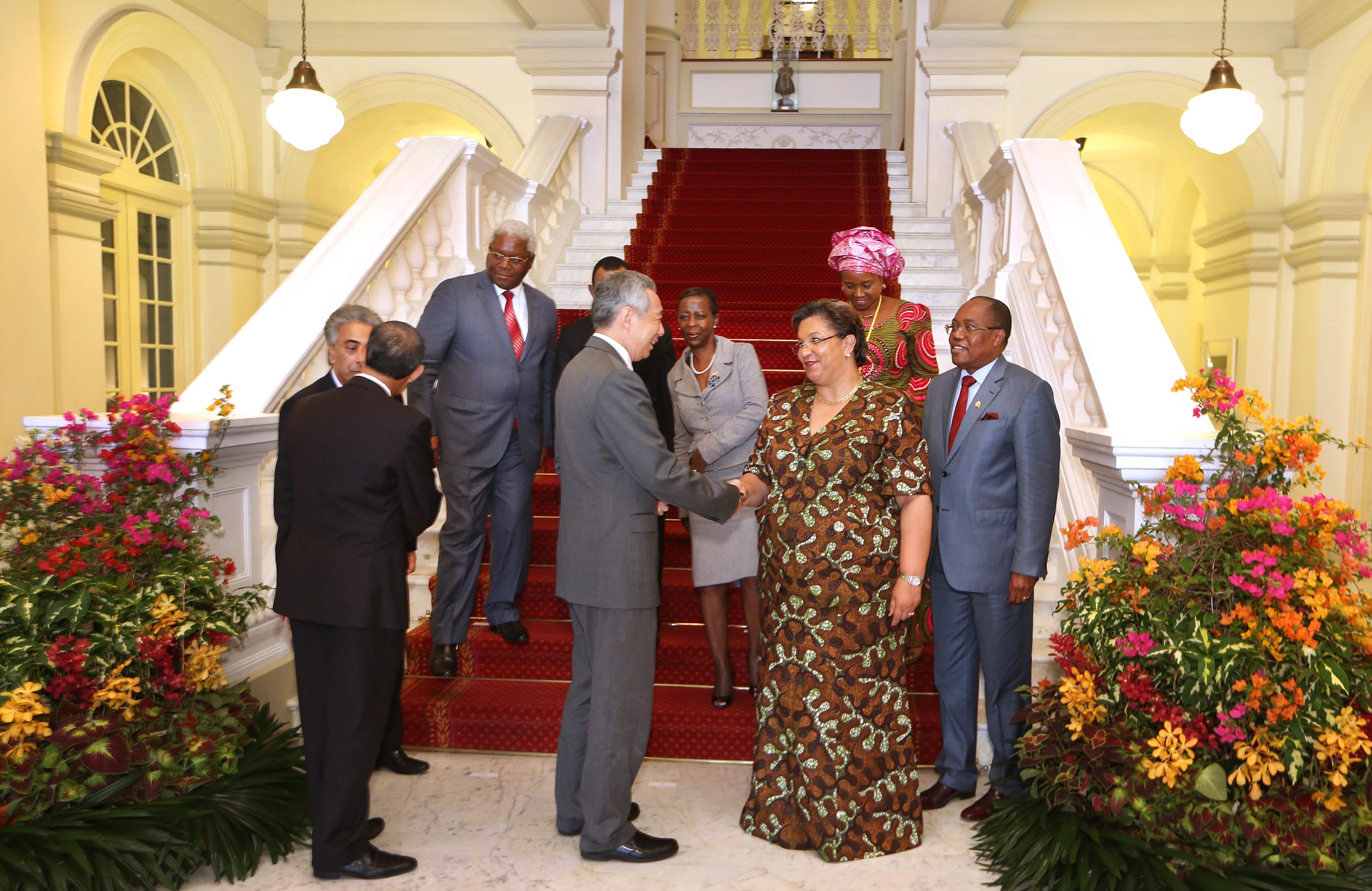 People in suits and dresses on staircase with red carpet, shaking hands, floral arrangements.