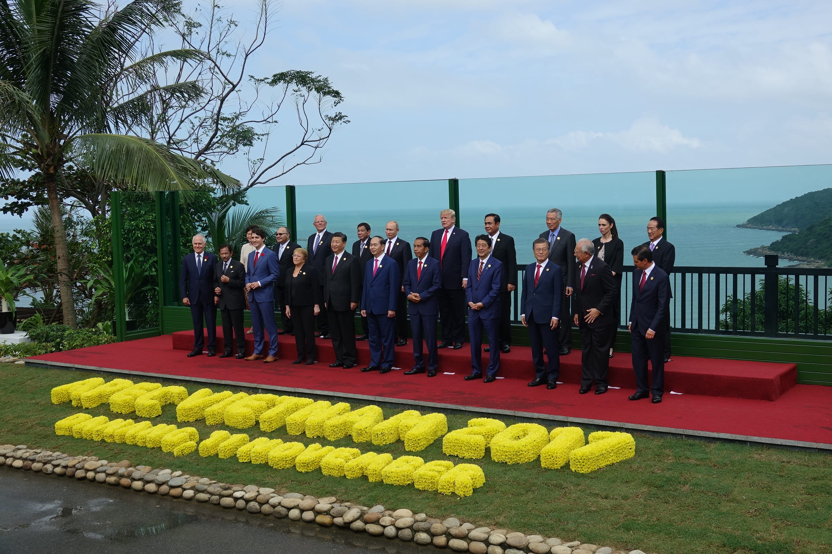 Group of leaders on red platform, sea behind, yellow flowers spell out letters.