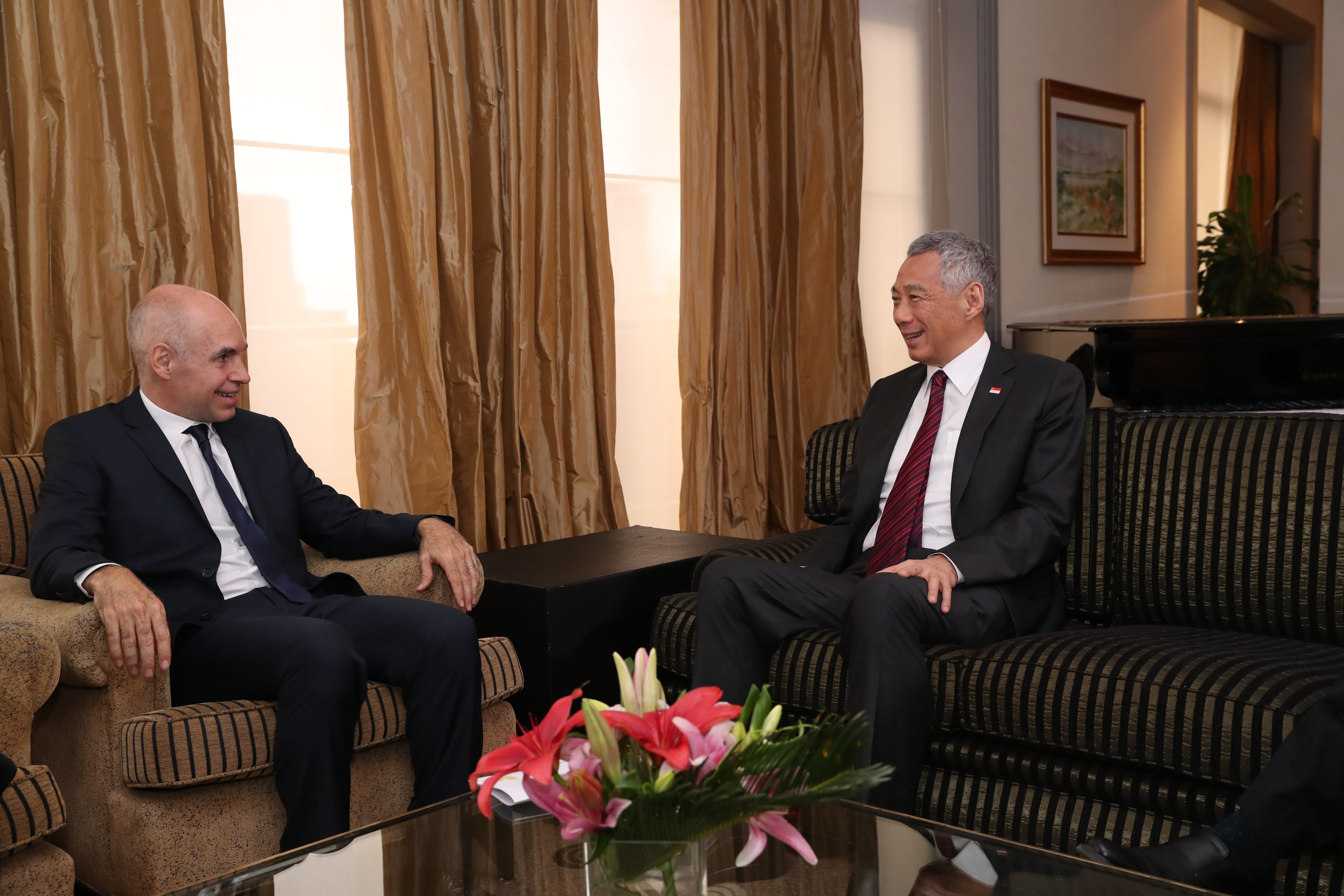 Two men in suits seated, facing each other in upholstered chairs, with a flower arrangement on a glass table.
