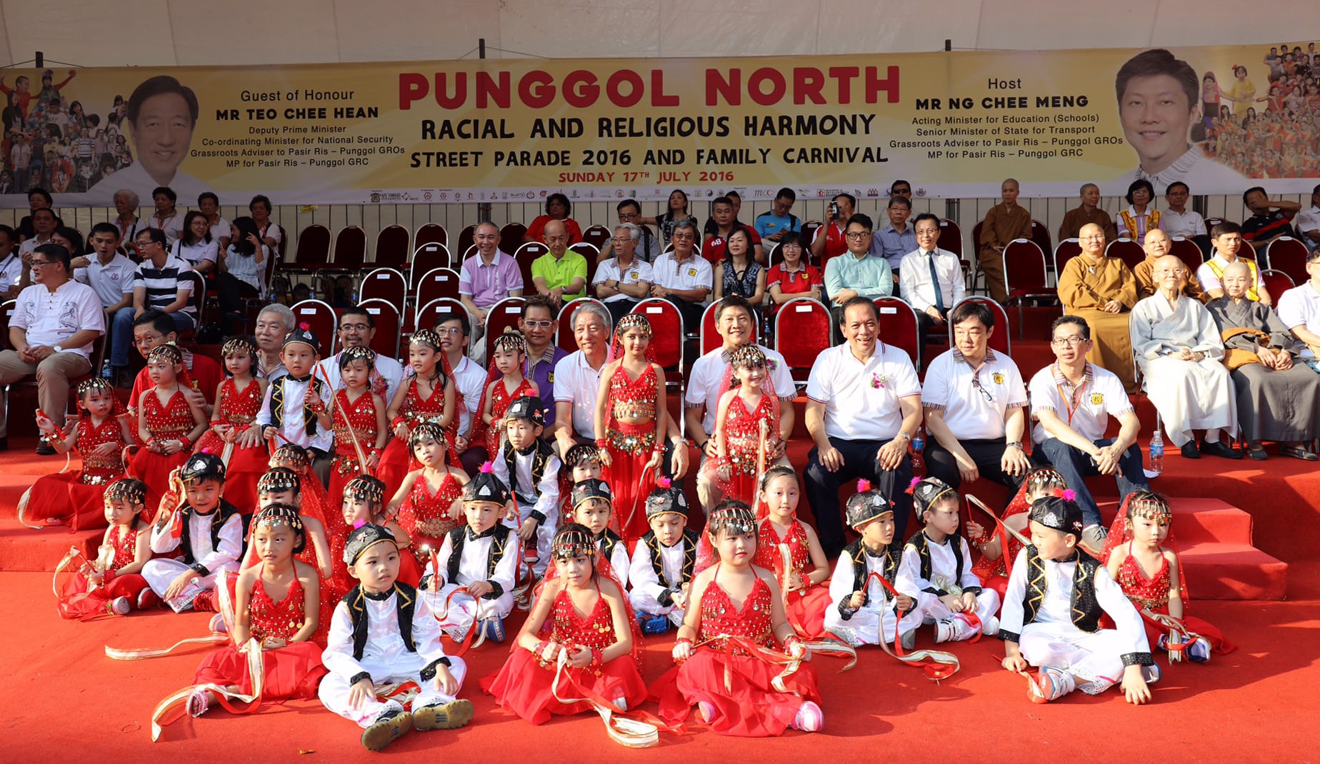 Group on red stage: children in cultural costumes, adults in background at Punggol North event.