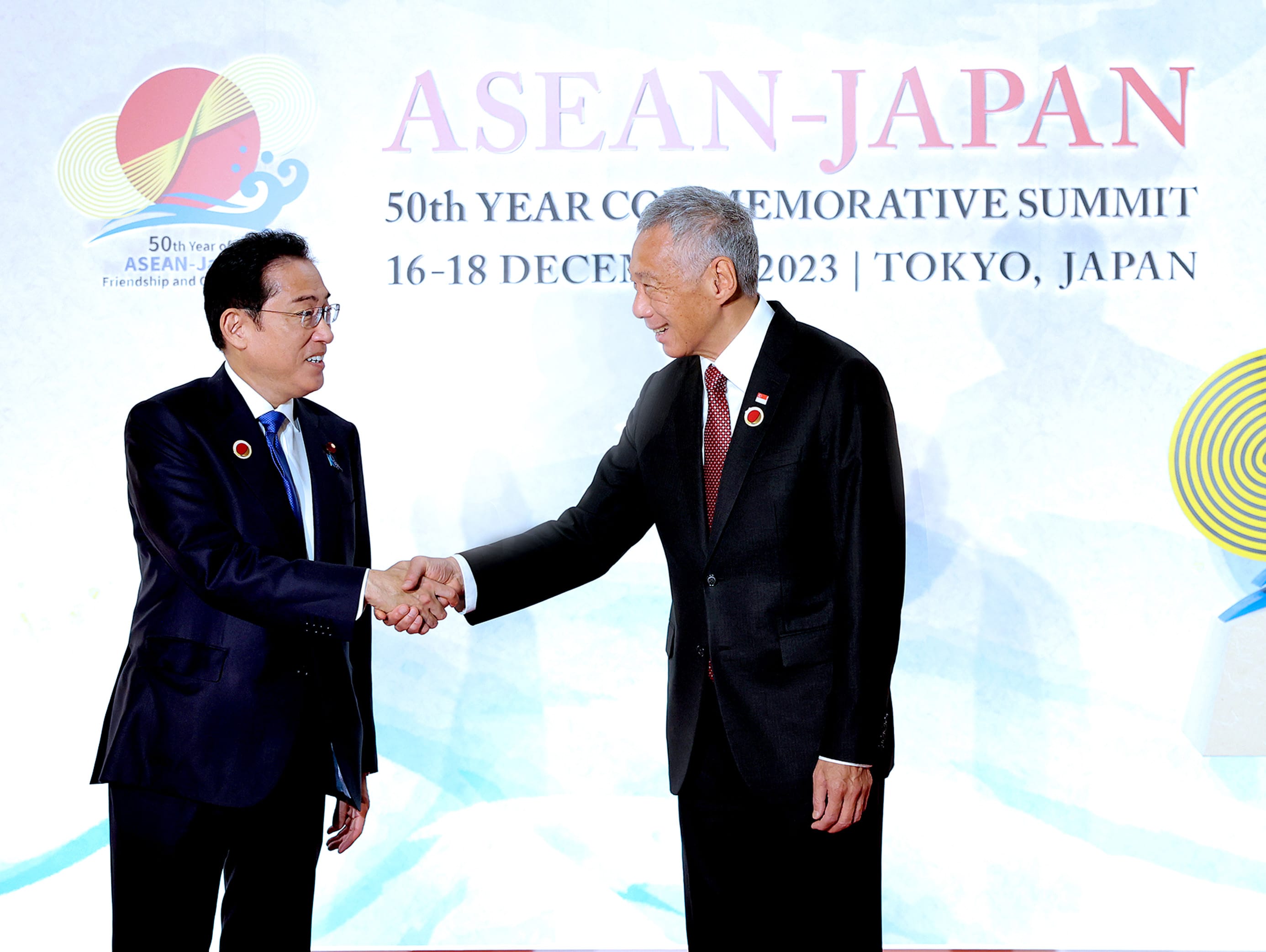 Two men in suits, one with a Japan lapel pin, shake hands in front of "ASEAN-JAPAN" banner.
