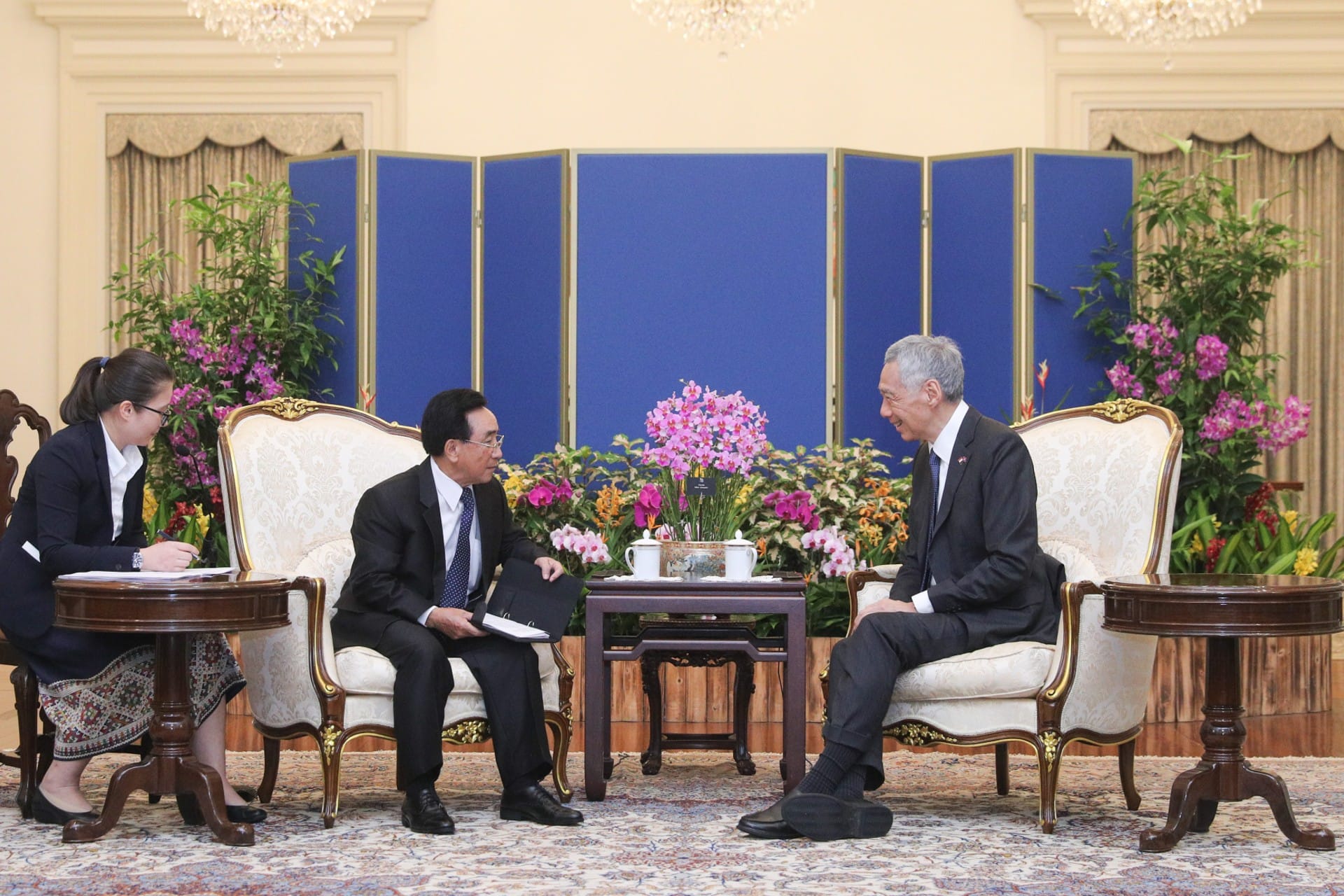 Two men in suits seated in ornate chairs facing each other, with a woman taking notes to the side.