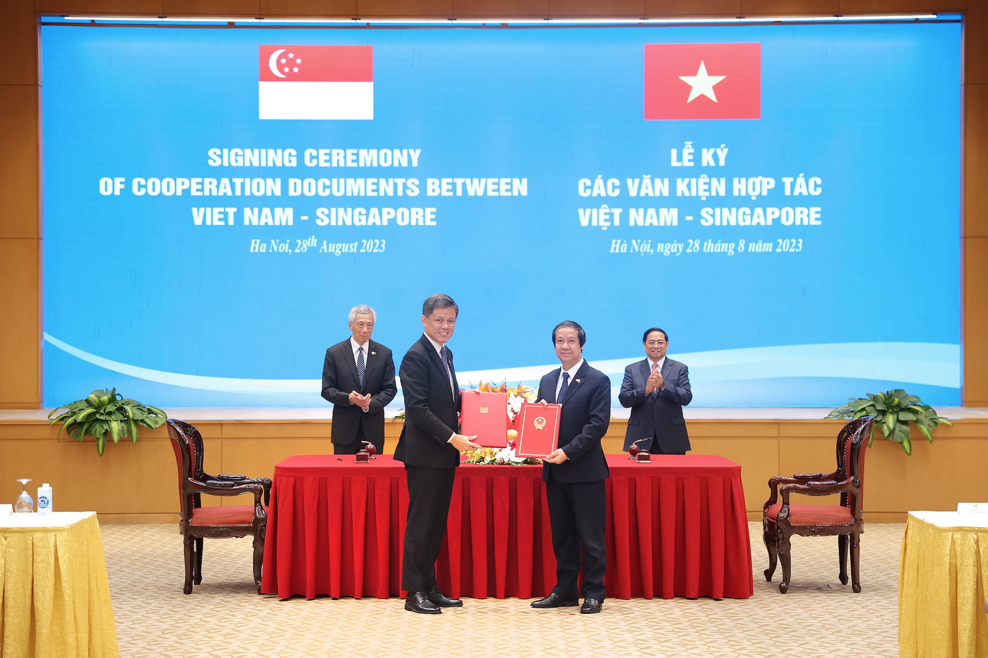 Two officials exchange documents at Viet Nam-Singapore signing ceremony, two men stand in background.