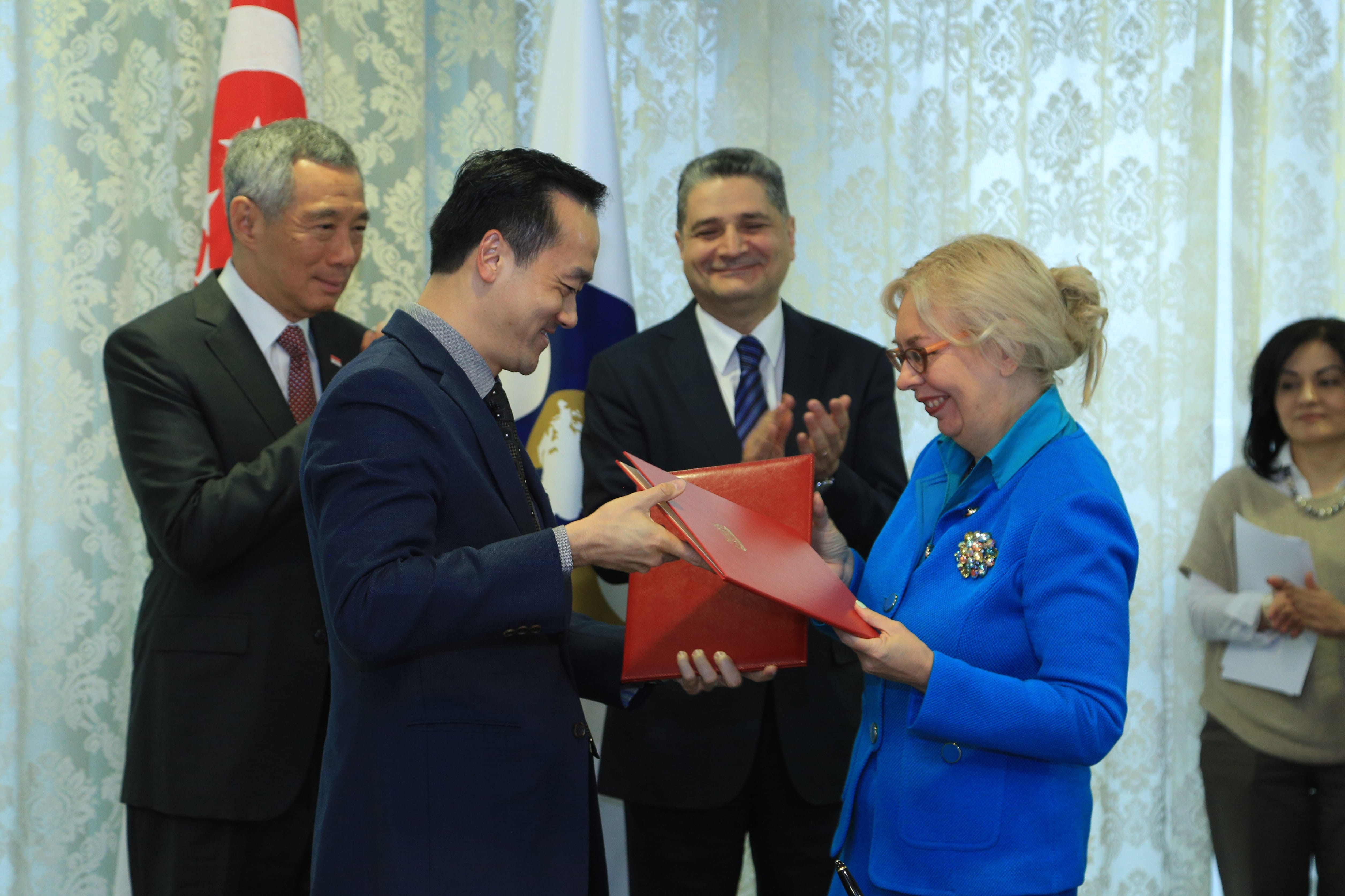 Four people near flags, one person handing a red folder to another wearing a blue suit.