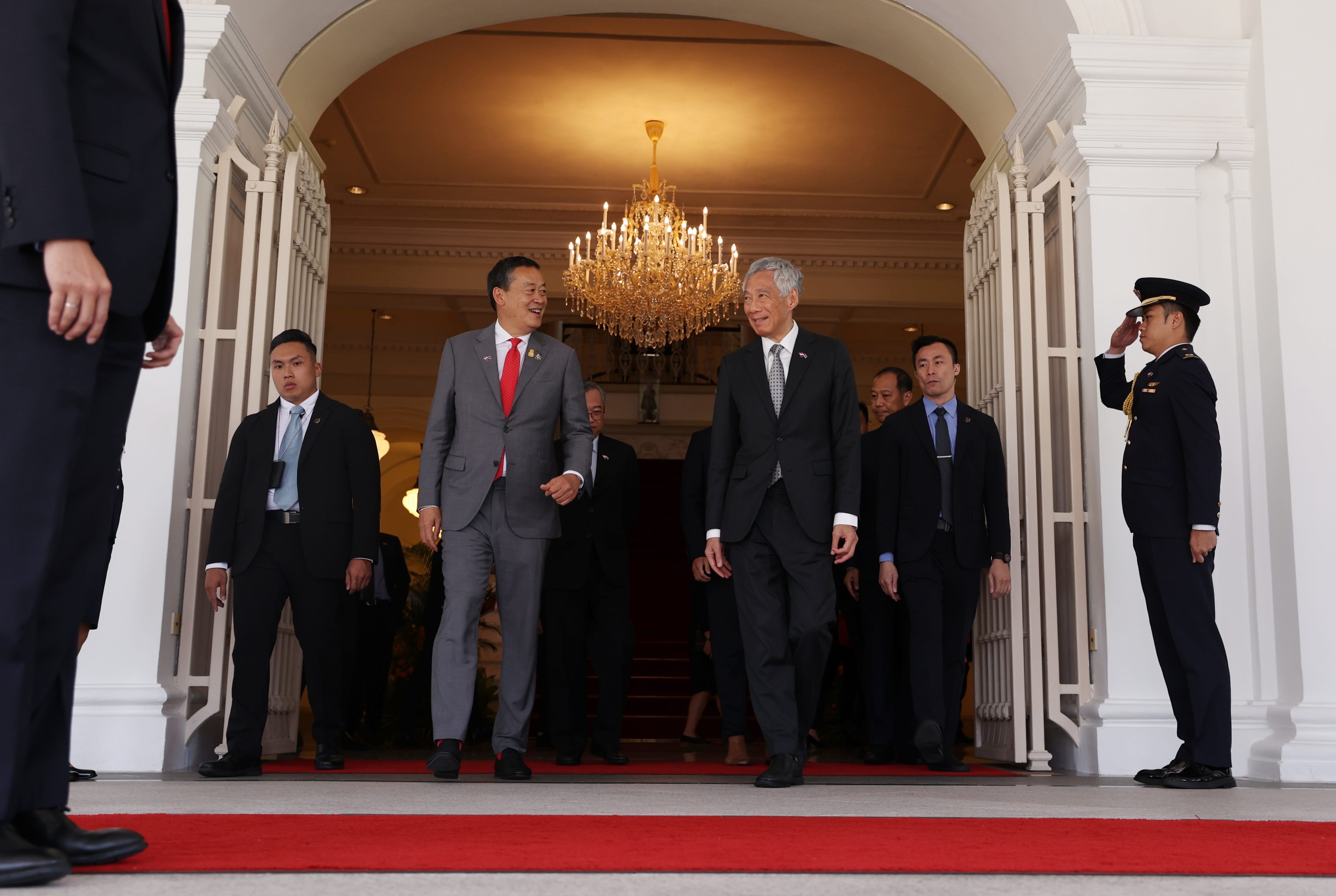 Lee Hsien Loong and another man in suits exit a building. A uniformed officer salutes. Red carpet.