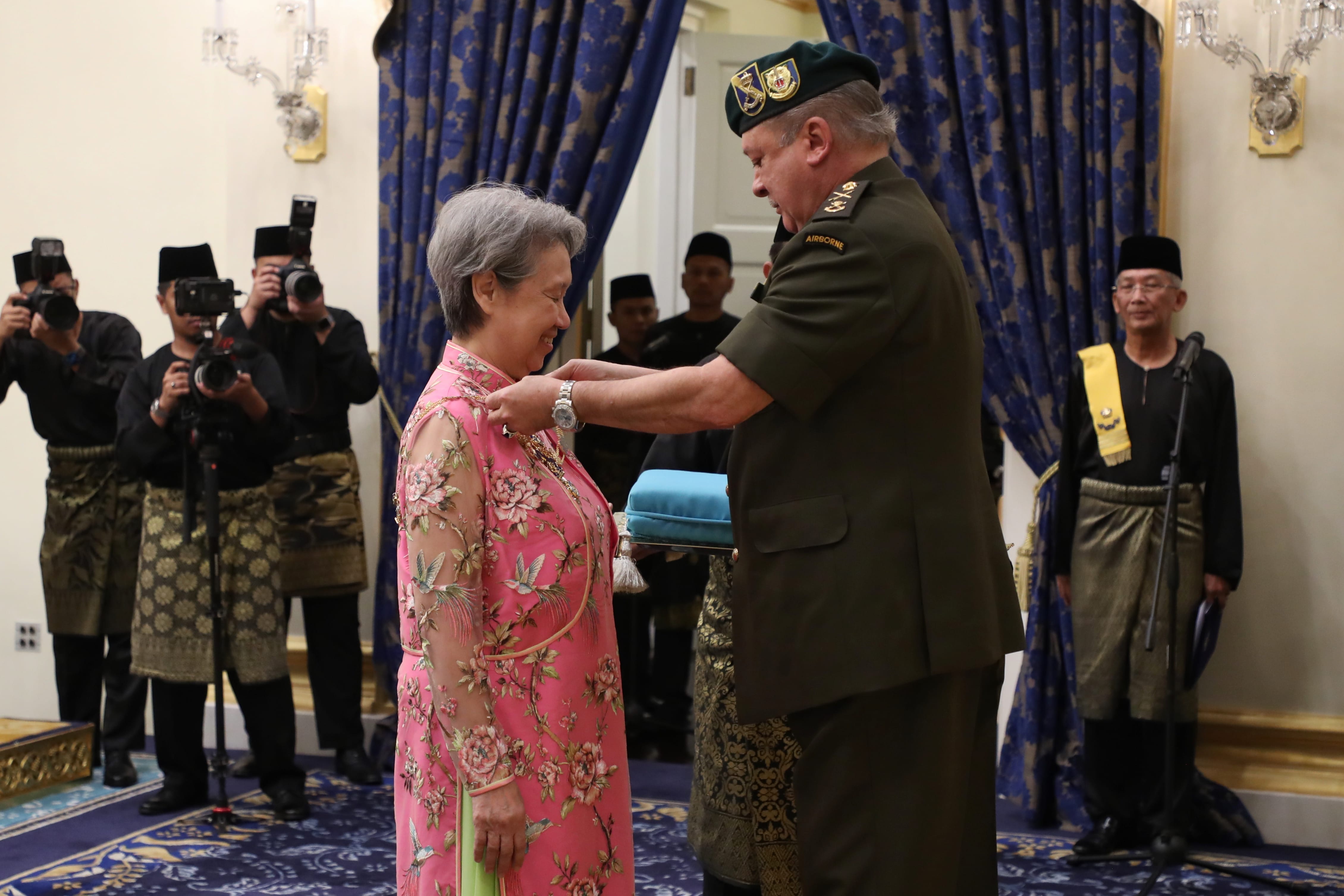 Man in military uniform pinning medal on woman in pink floral dress. Photographers in background.