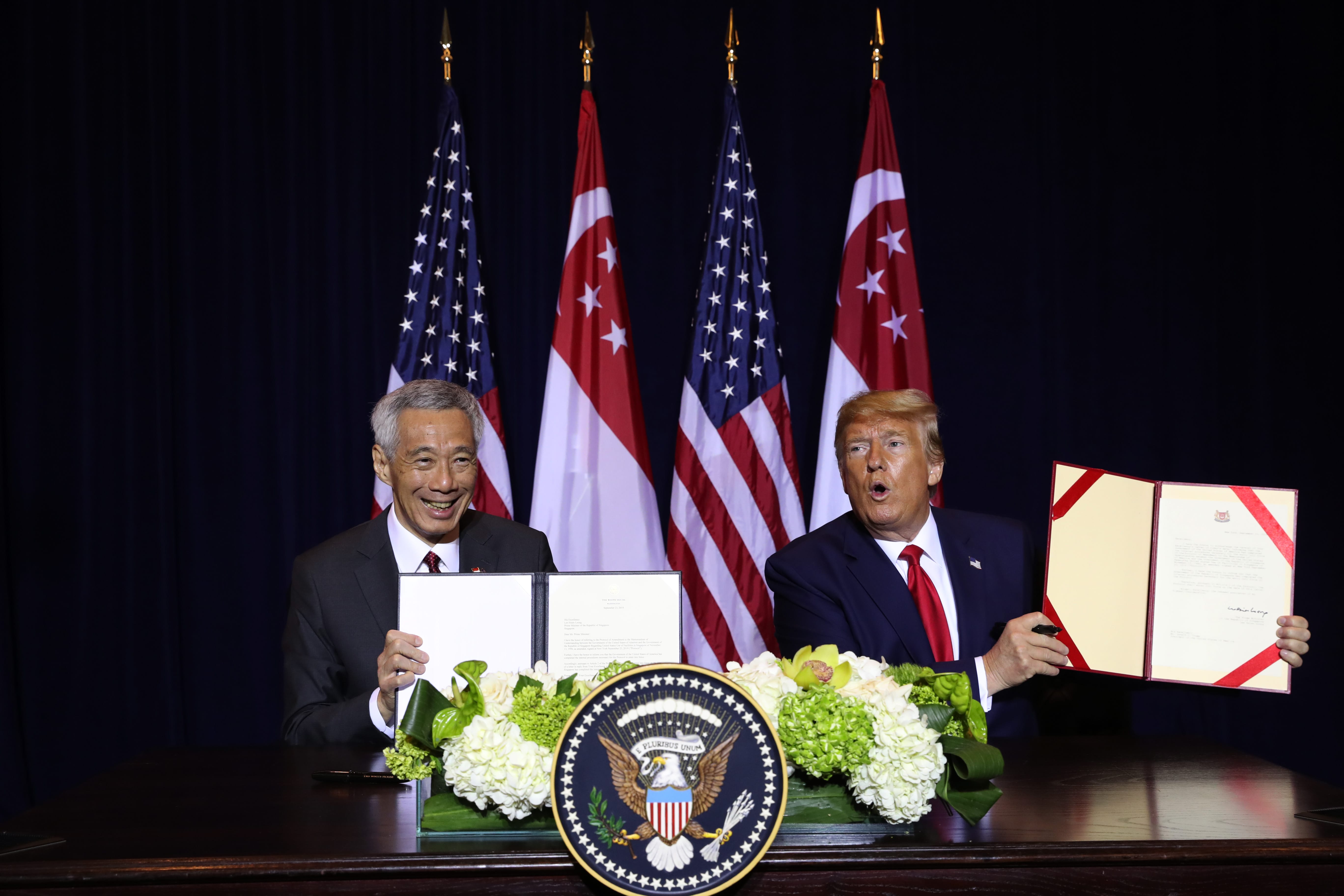Lee Hsien Loong and Donald Trump hold documents with US and Singapore flags in the background.