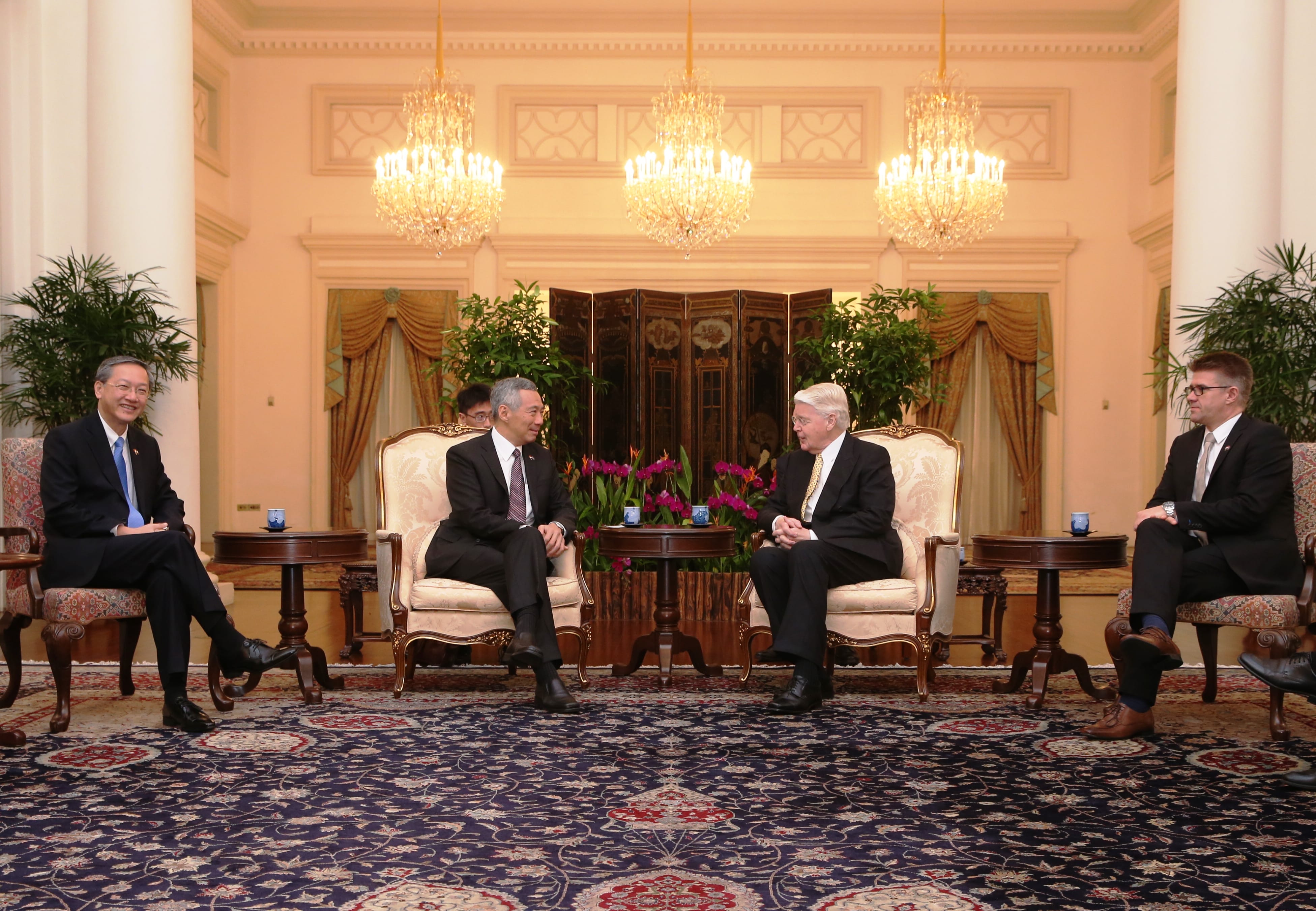 Four people in suits sit in armchairs, evenly spaced, on ornate rug in a room with chandeliers.
