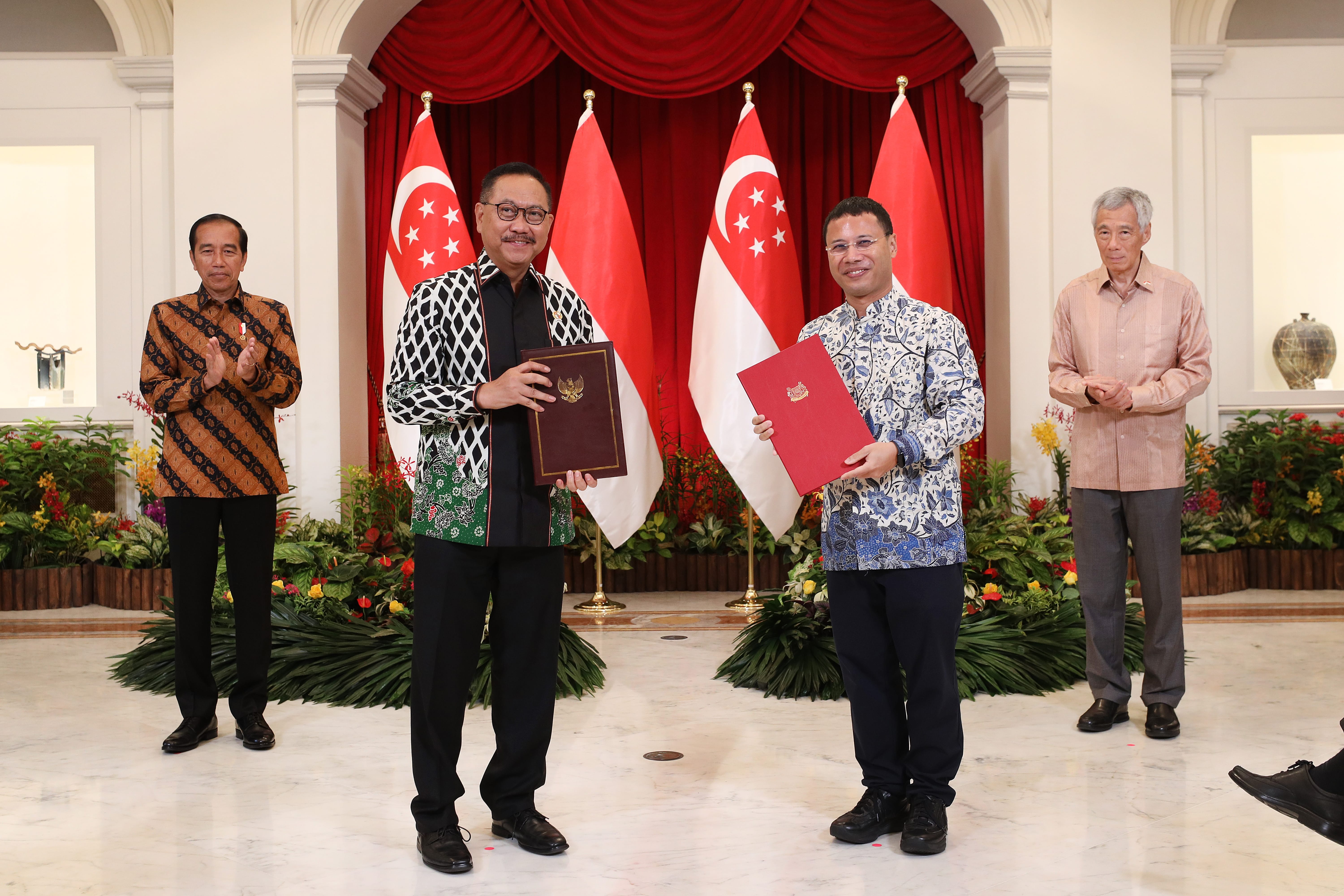 Four men in suits hold documents before Singaporean flags in a room.