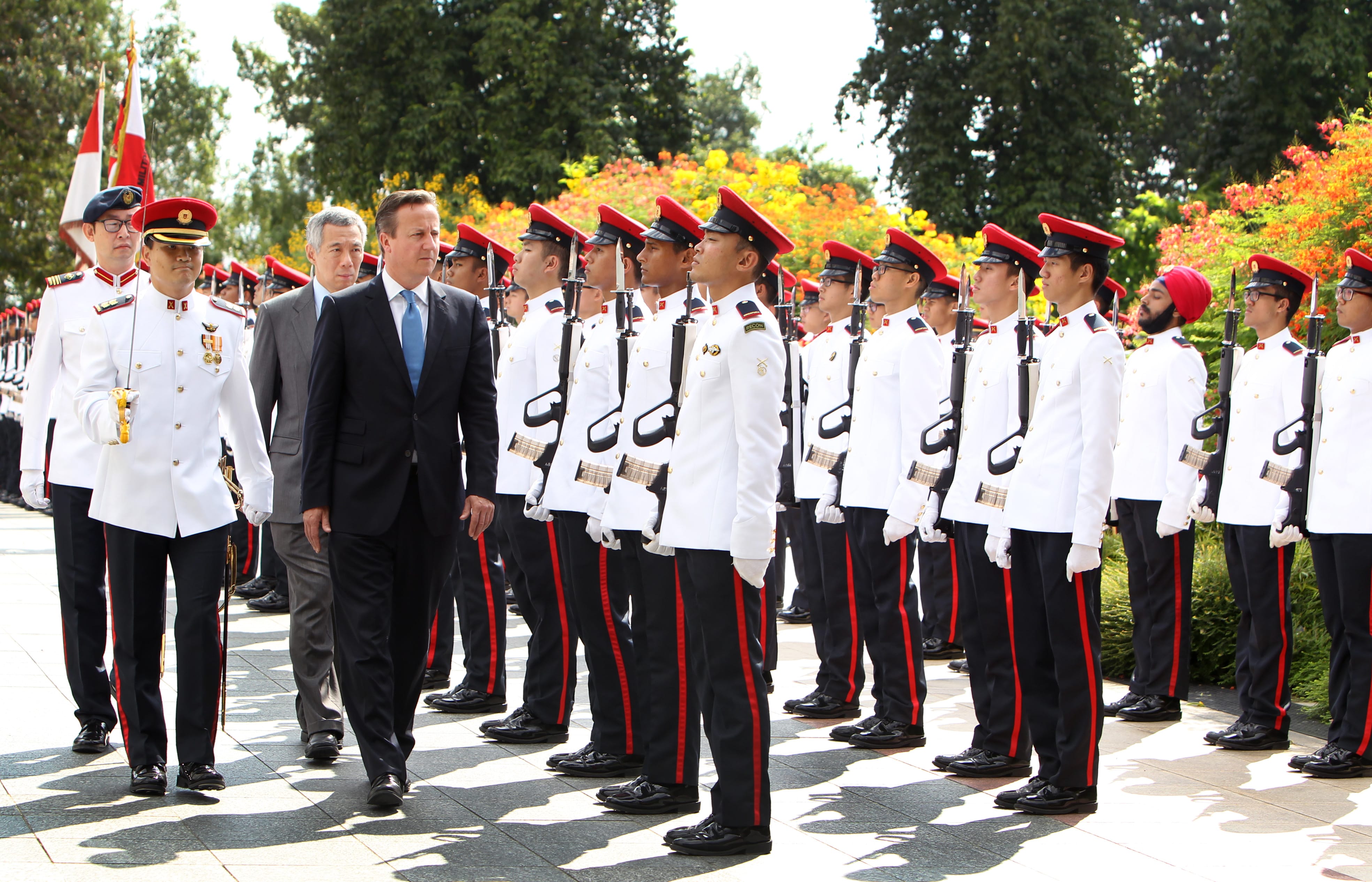 David Cameron walks past soldiers in white uniforms with red trim, holding rifles.