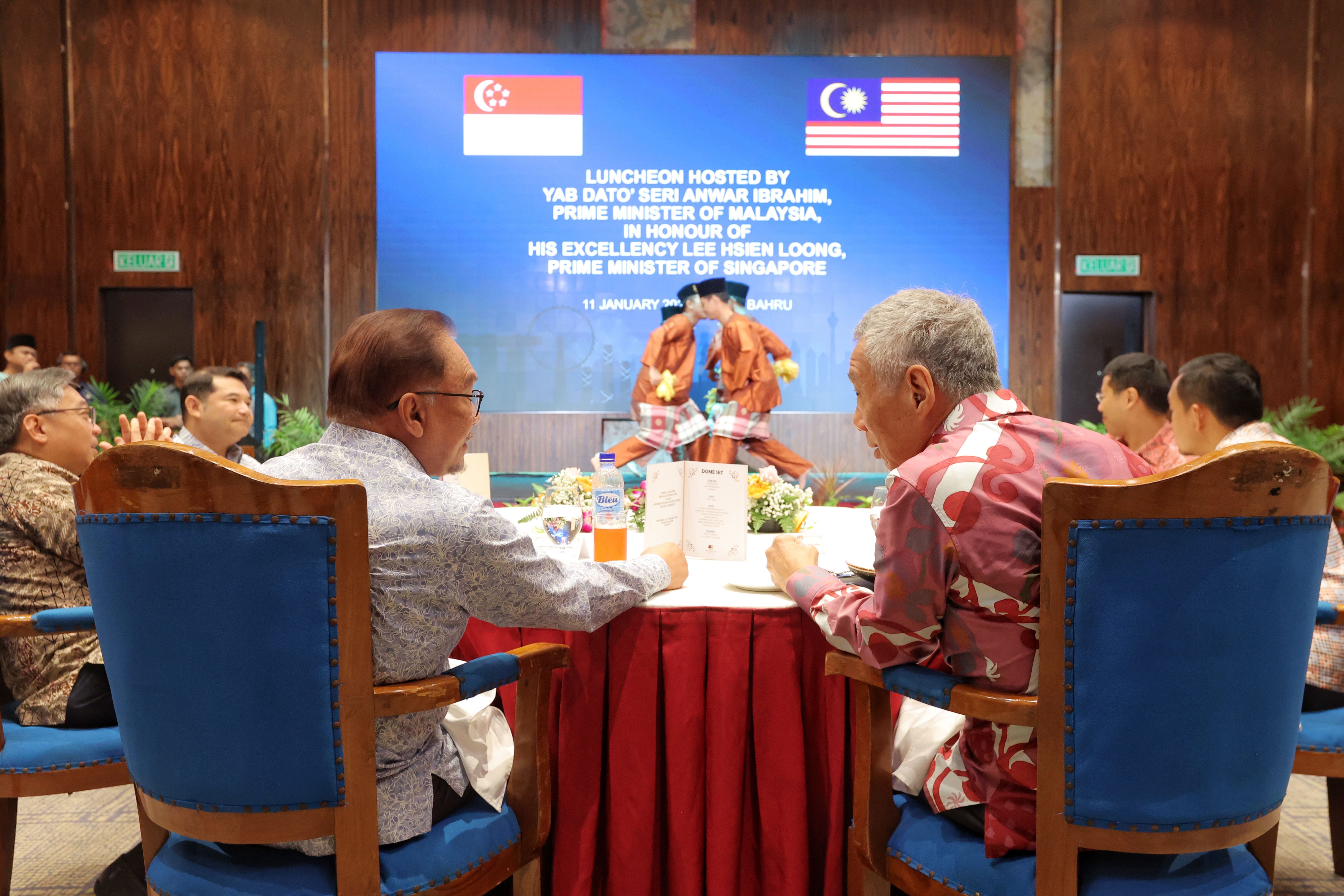 Two men seated at a table; stage backdrop with flags of Singapore and Malaysia.