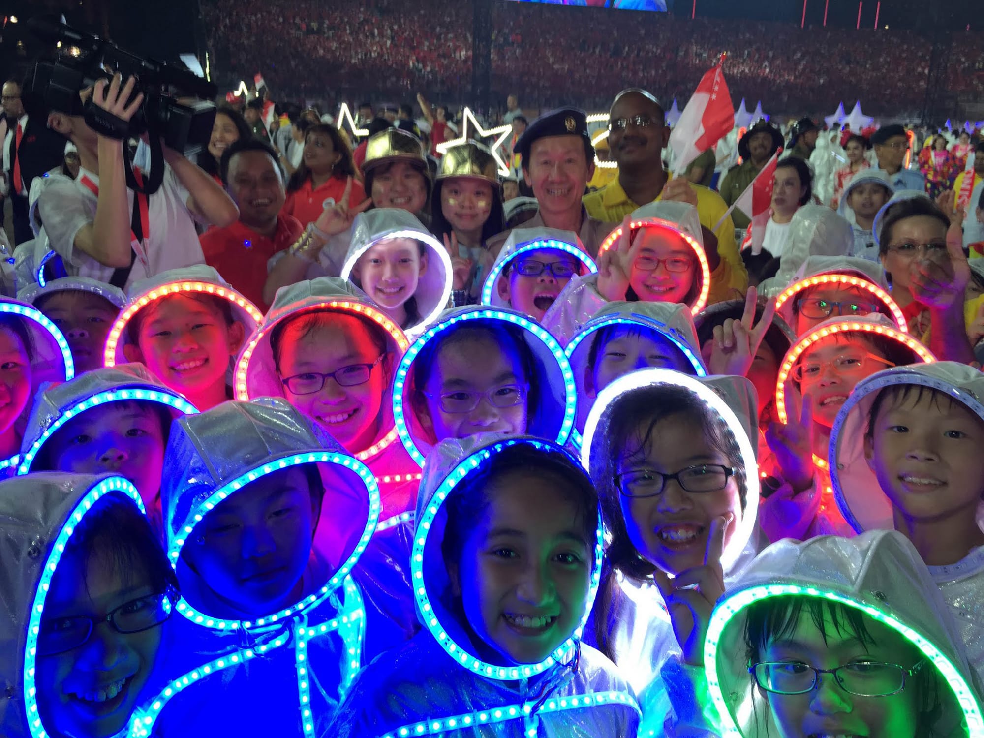 Crowd of children in lit costumes and adults, some with Singapore flags, at event.
