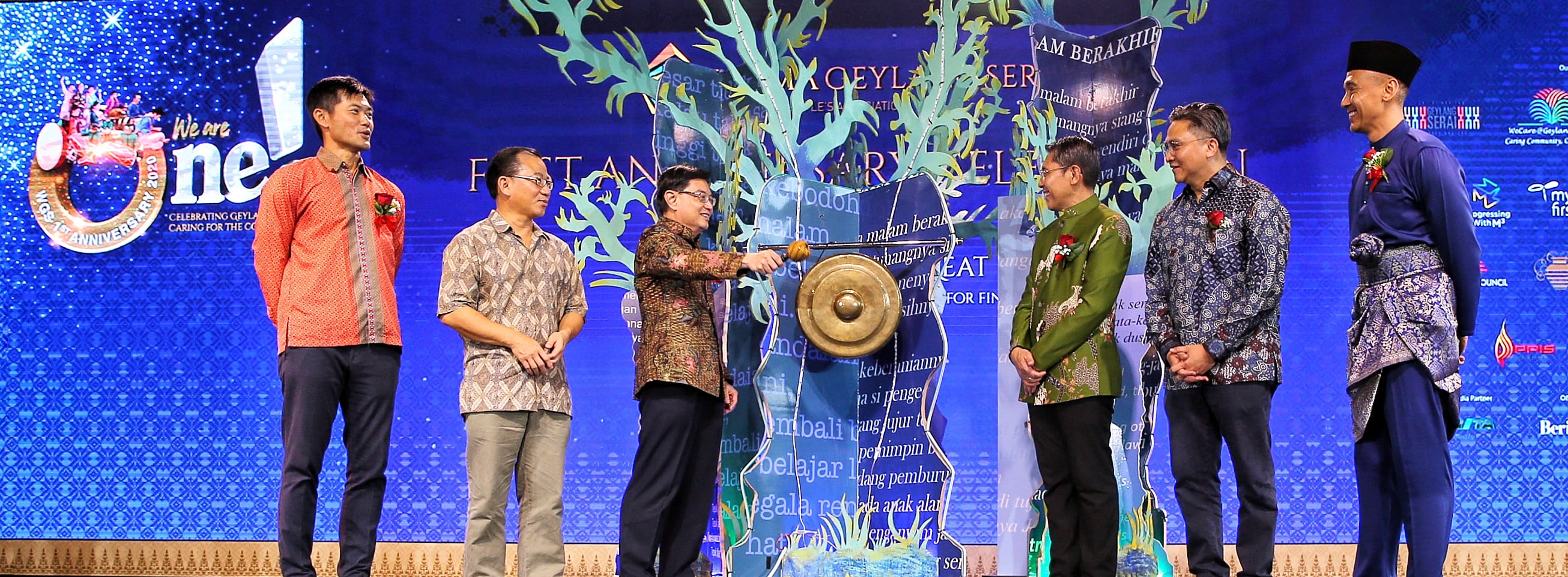 Six people in formal wear stand on a stage, one striking a gong in front of a decorative screen.