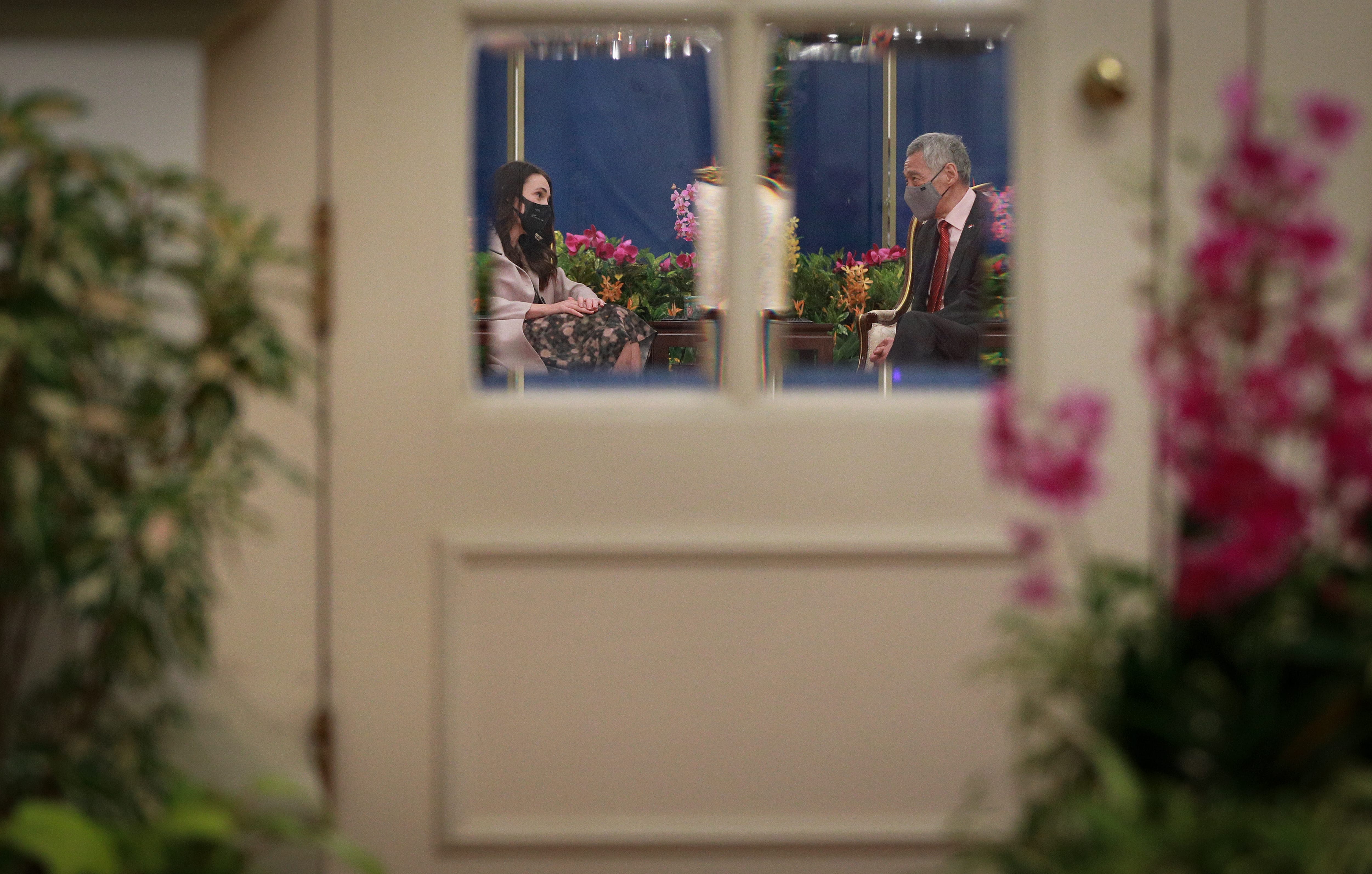 Through doorway, Jacinda Ardern and Lee Hsien Loong wearing masks, seated amongst floral decor.