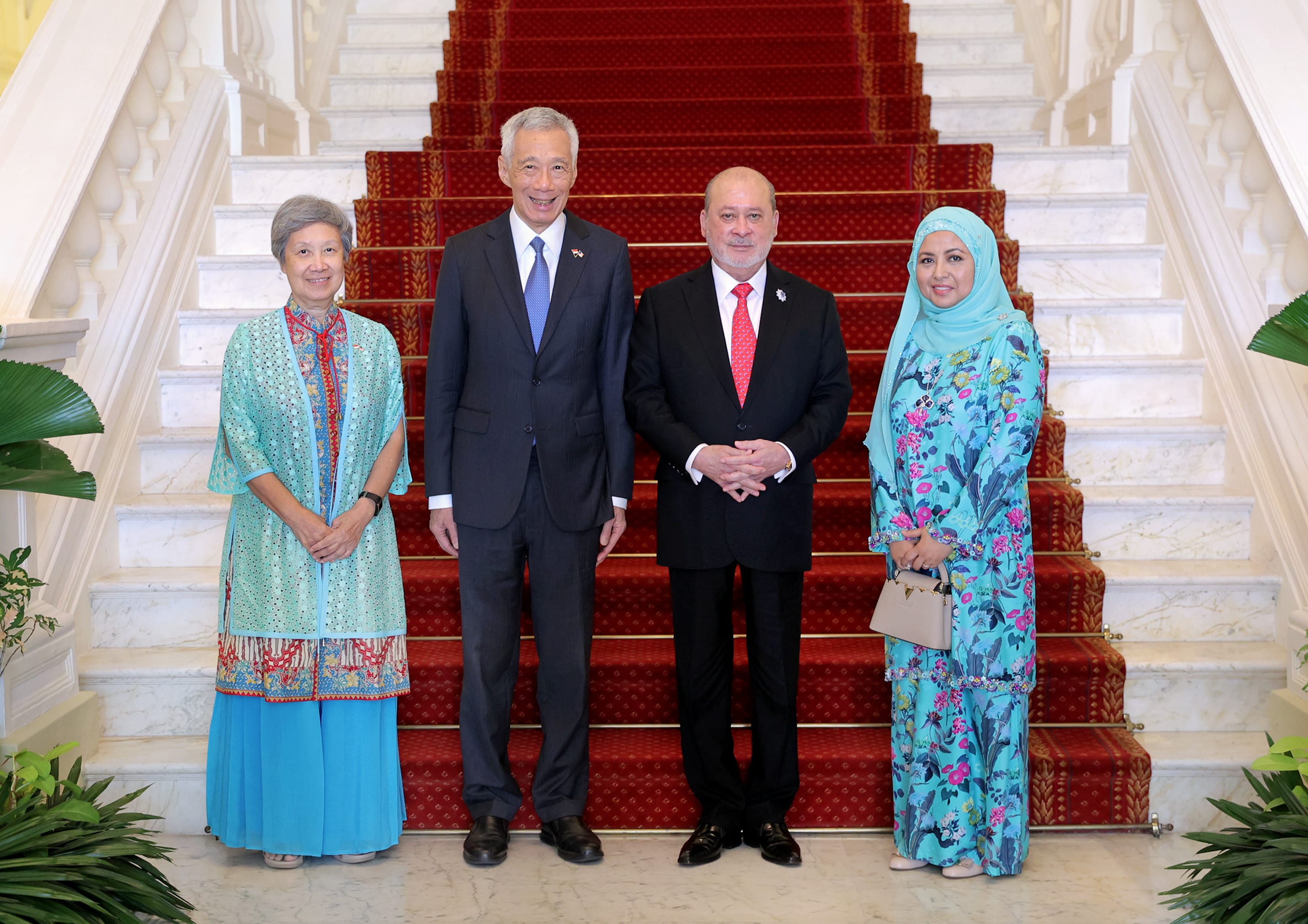 Four people on stairs covered in red patterned carpet. Two men in suits, two women in patterned outfits.