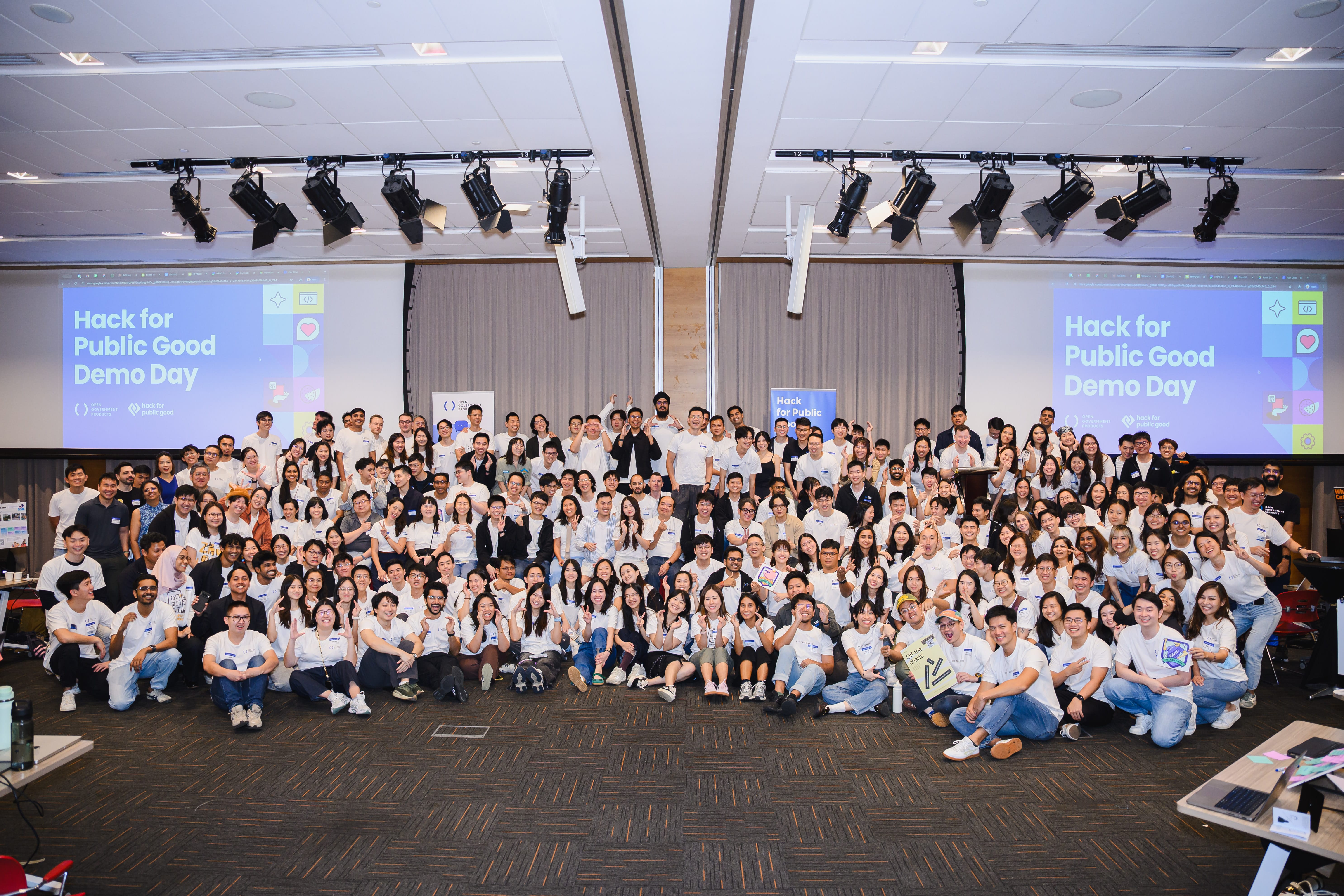 Large group photo of community hackathon participants posing together in an event hall with QR codes on screens