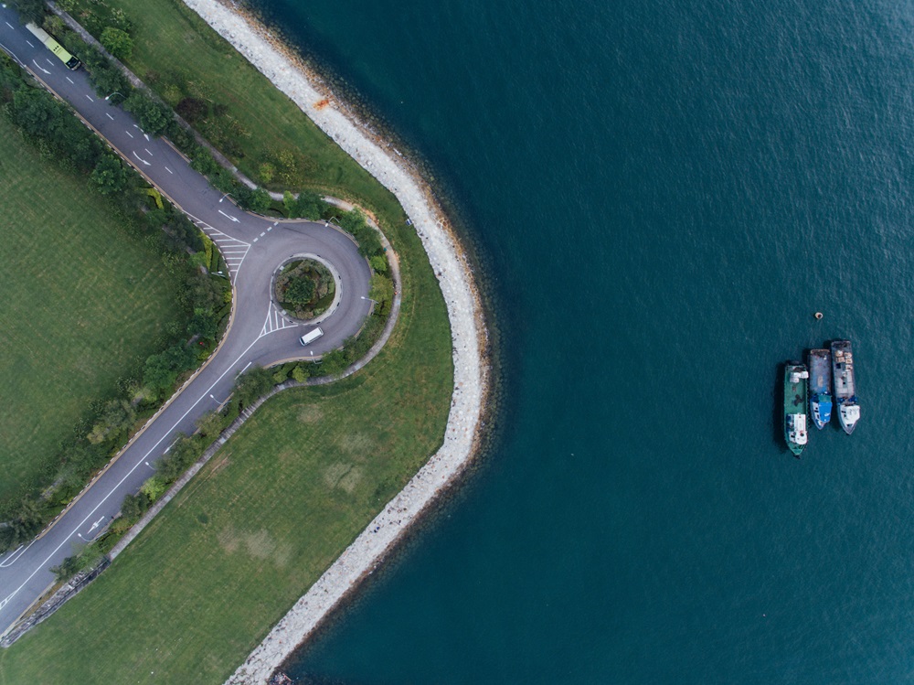 Aerial view of a roundabout on a grassy peninsula by a rocky shoreline, with three boats floating in blue water.