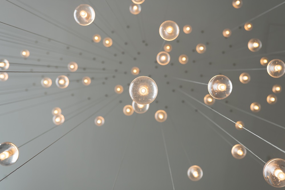 Suspended art installation of dozens of glowing bulbs hanging at varied heights against a gray ceiling.